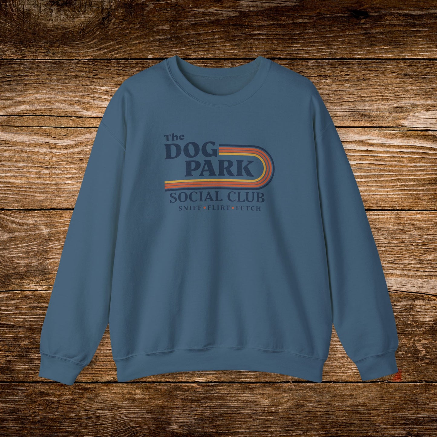 A slate blue crewneck sweatshirt featuring a retro-style graphic that reads 'The DOG PARK SOCIAL CLUB' with 'SNIPER, PLOYER, FETCH' underneath, displayed flat against a rustic brown wooden plank background. The design text reads "Sniff, Flirt, Fetch."