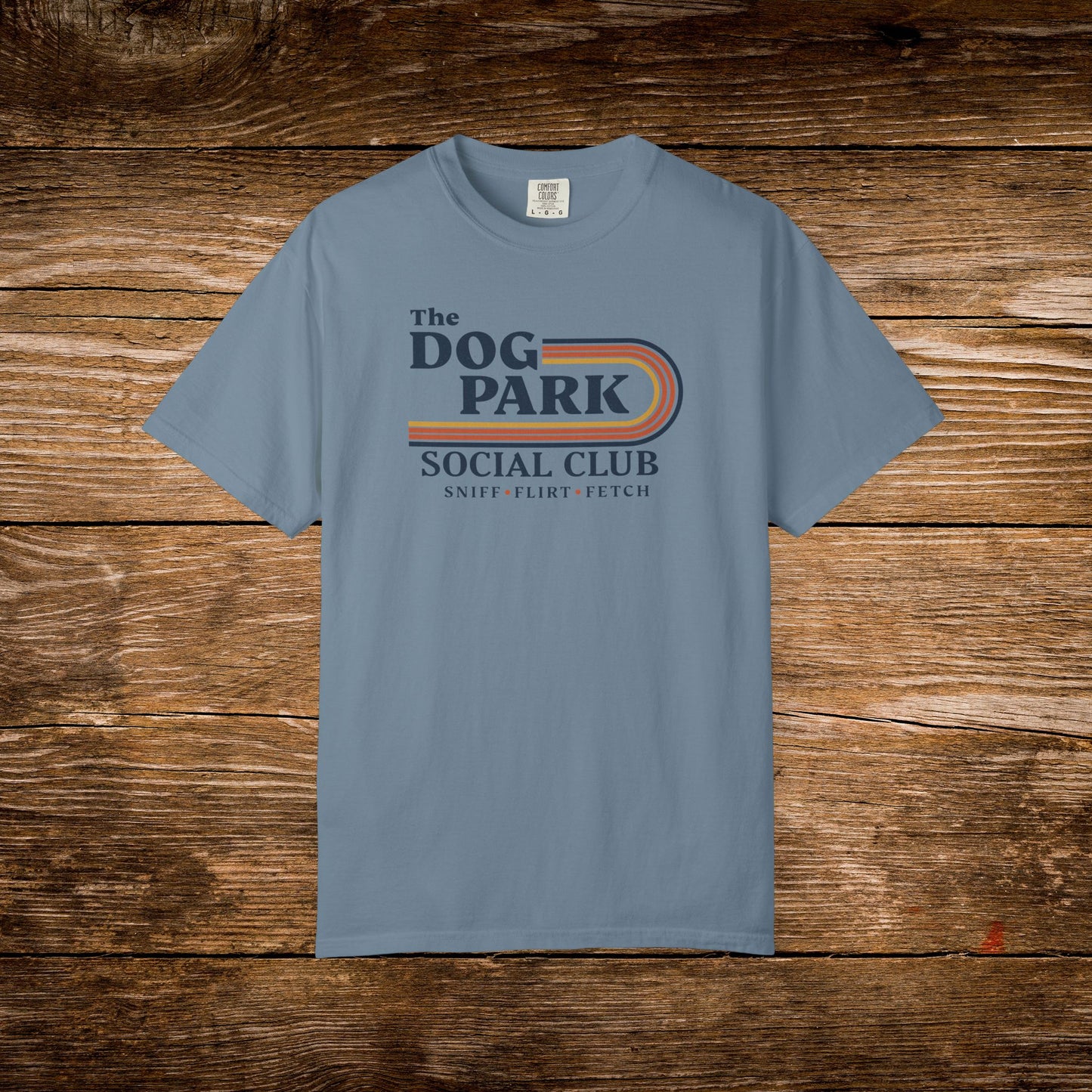A light blue crewneck t-shirt with a retro-style graphic reading 'The Dog Park Social Club' and 'Sniff, Flirt, Fetch' in navy and orange, displayed flat against a rustic brown wood plank background.