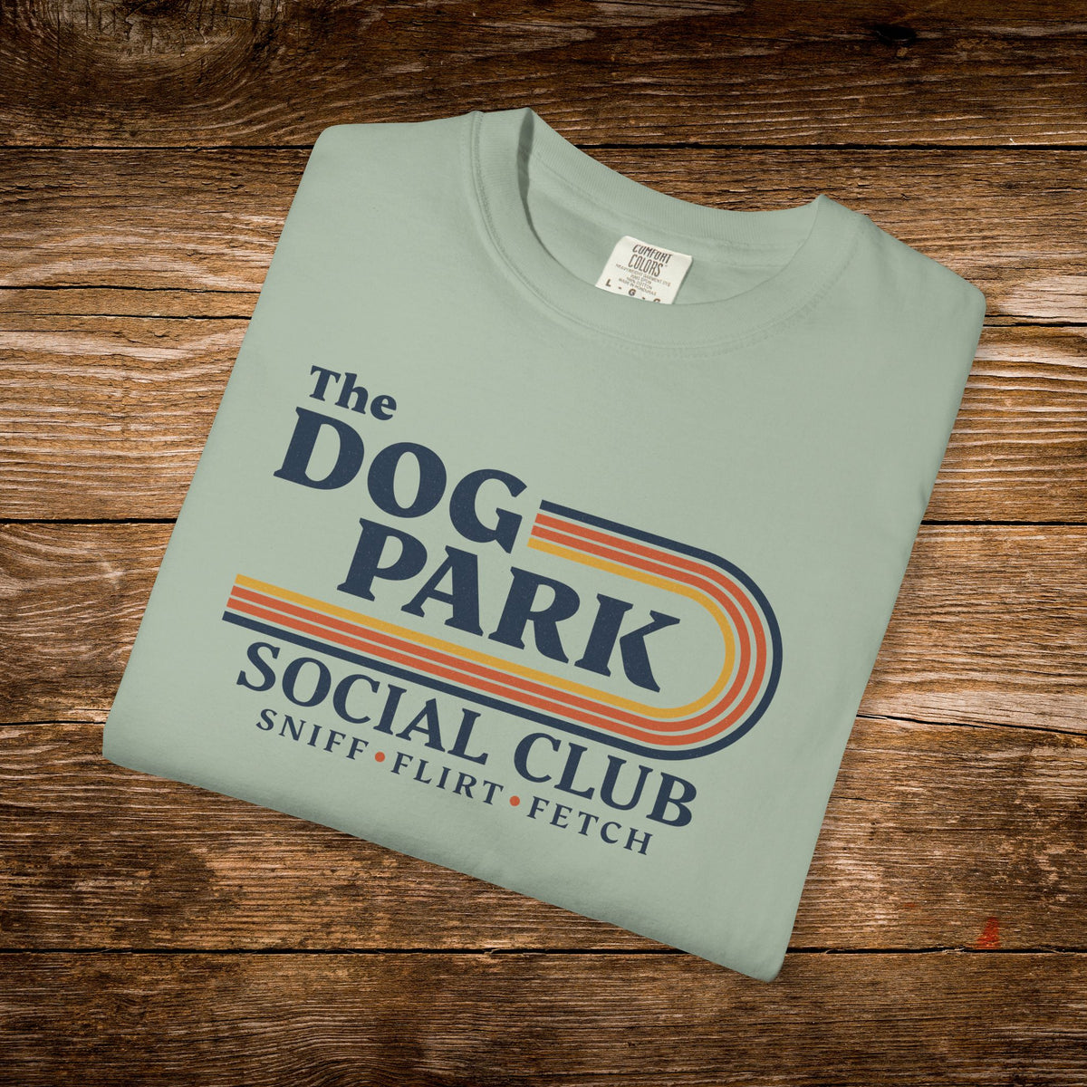 A folded sage green t-shirt with a retro-style graphic reading 'The DOG PARK SOCIAL CLUB' and 'SNIFF • FLIRT • FETCH' in navy, orange, and yellow, displayed on a rustic wooden background.