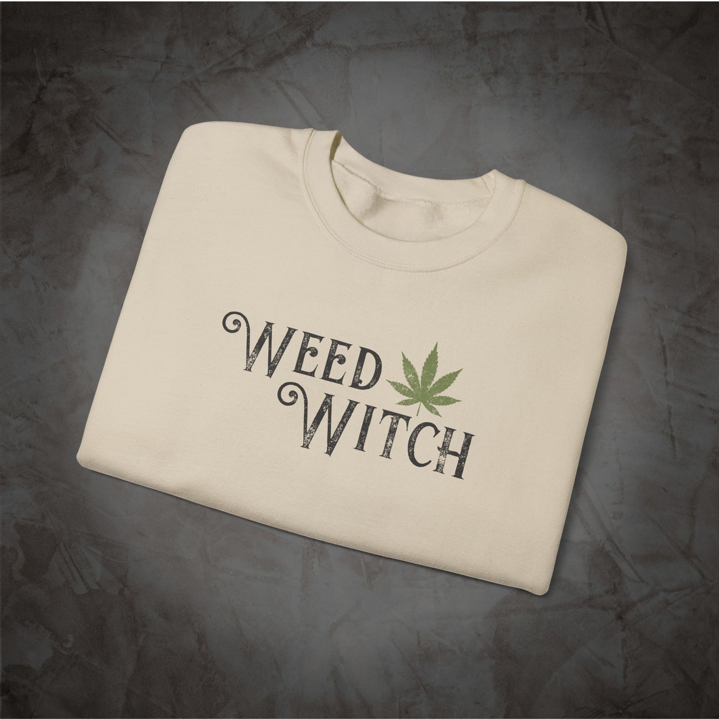 An off-white crewneck sweatshirt with 'WEED WITCH' printed in a dark gothic font and a green cannabis leaf graphic, laid flat on a dark textured background.