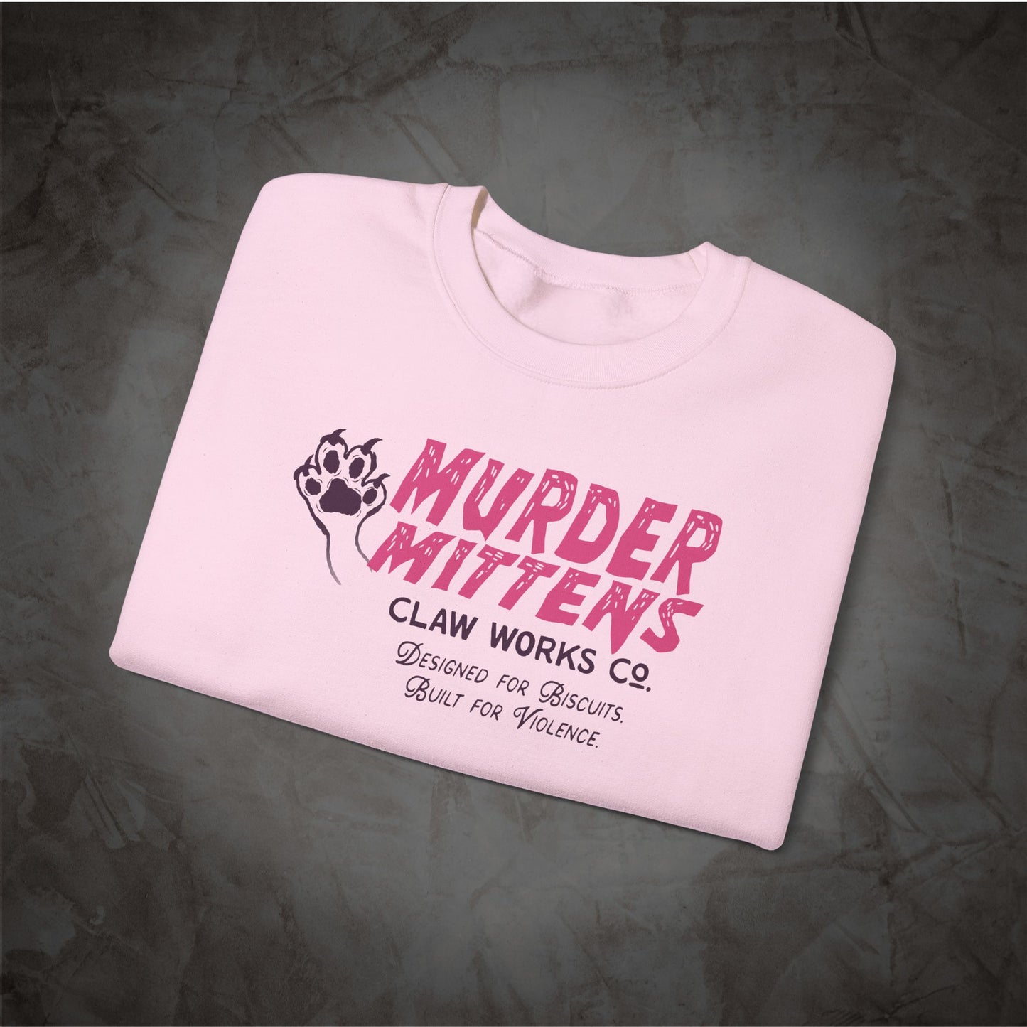 A folded light pink t-shirt with a graphic print reading 'MURDER MITTENS' in distressed pink letters, alongside a black paw print logo and smaller text 'CLAW WORKS CO. Designed for Biscuits.