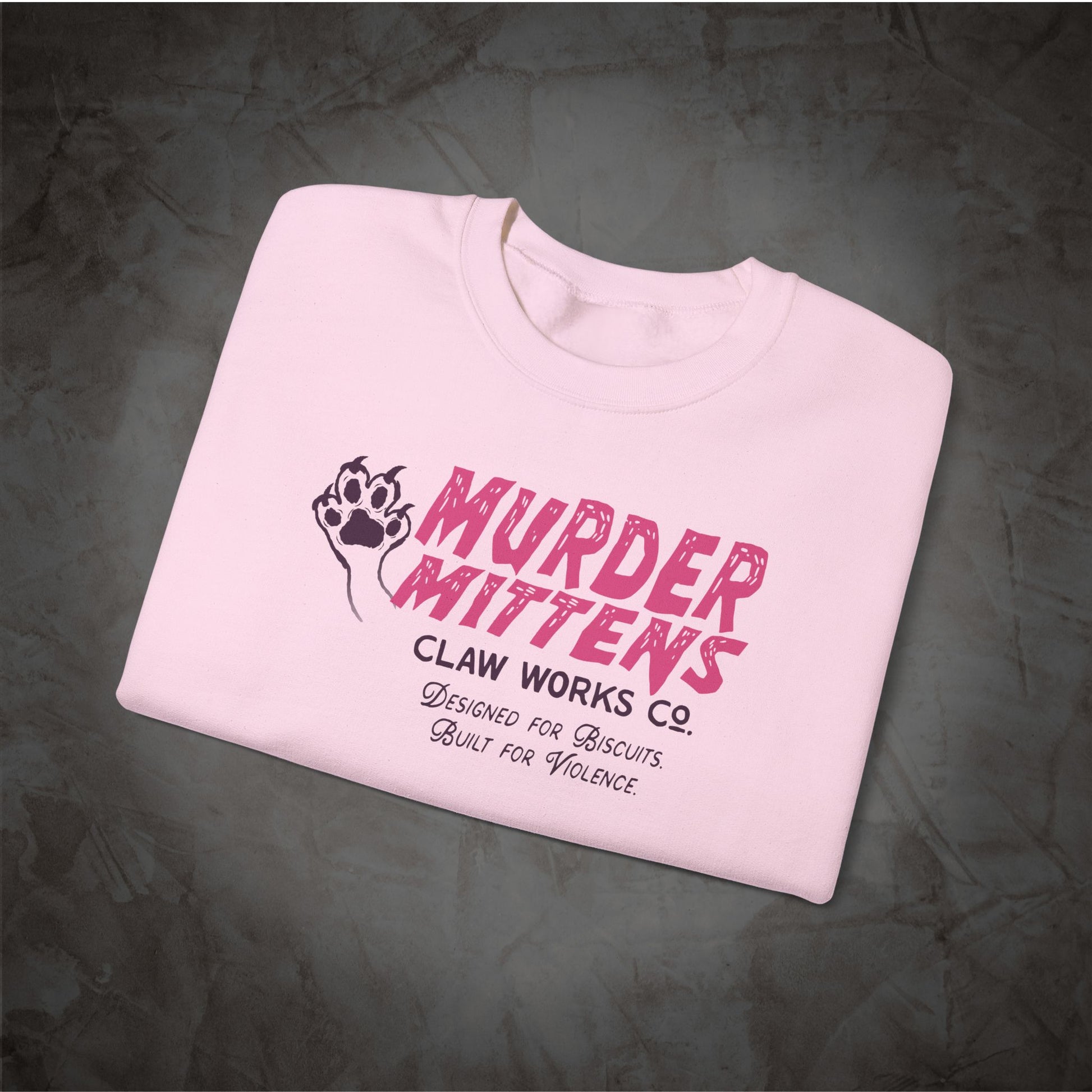 A folded light pink t-shirt with a graphic print reading 'MURDER MITTENS' in distressed pink letters, alongside a black paw print logo and smaller text 'CLAW WORKS CO. Designed for Biscuits.