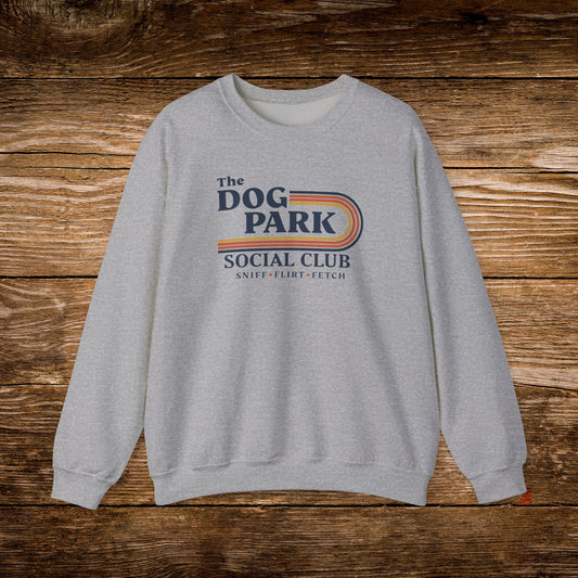 A light gray crewneck sweatshirt with a retro-style graphic reading 'The Dog Park Social Club' and 'Sniff, Flirt, Fetch' in navy and orange, displayed flat against a rustic wooden plank background.