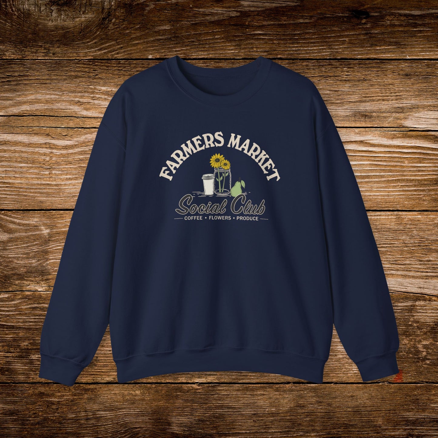 A navy blue crewneck sweatshirt with a vintage-style graphic on the chest reading 'FARMERS MARKET Social Club' in cream and yellow, featuring a sunflower, coffee cup, and green leaves, displayed flat against a rustic brown wooden background.