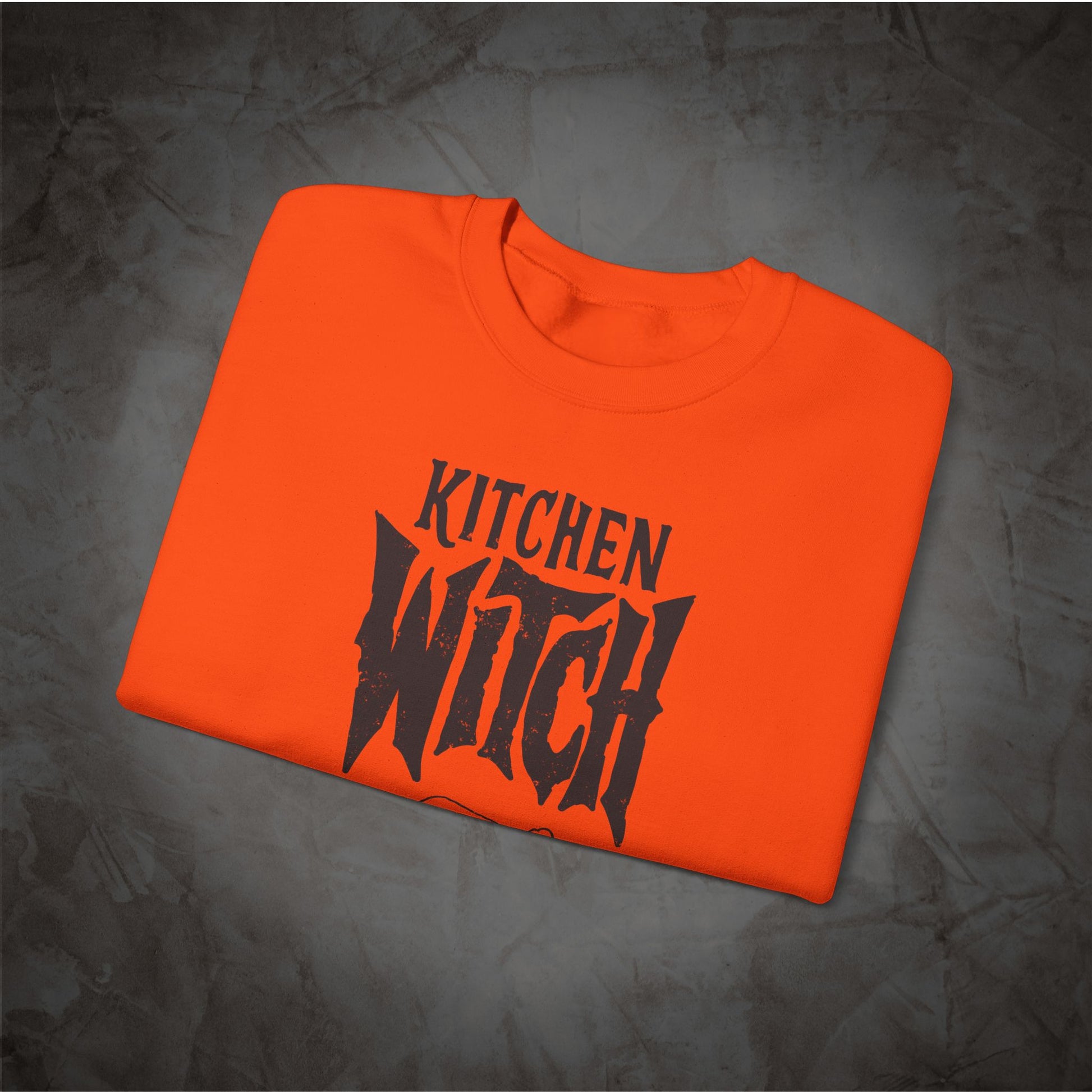 A bright orange folded t-shirt with a black graphic reading 'KITCHEN WITCH' in a stylized font, displayed on a textured dark gray background.