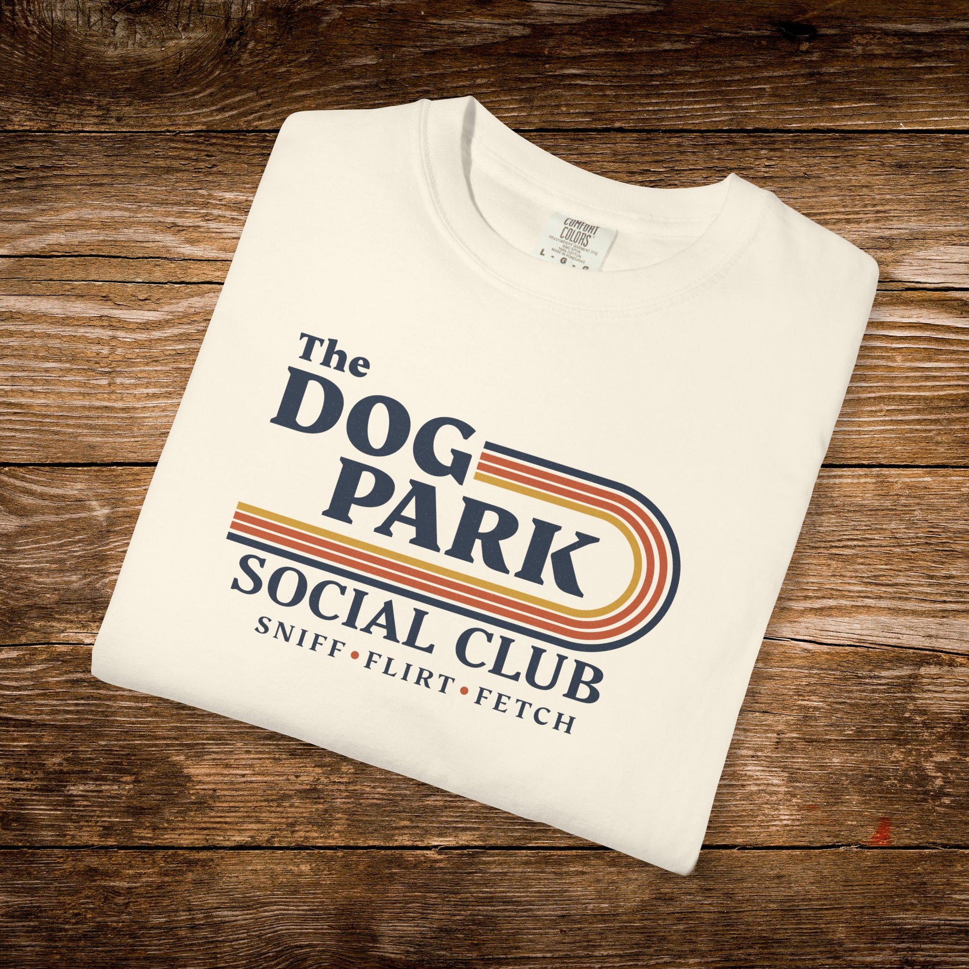 A folded off-white t-shirt with a retro-style graphic reading 'The DOG PARK SOCIAL CLUB' and 'SNIFF • FLIRT • FETCH' in navy, orange, and yellow, displayed on a rustic brown wooden background. The design text reads "Sniff, Flirt, Fetch."