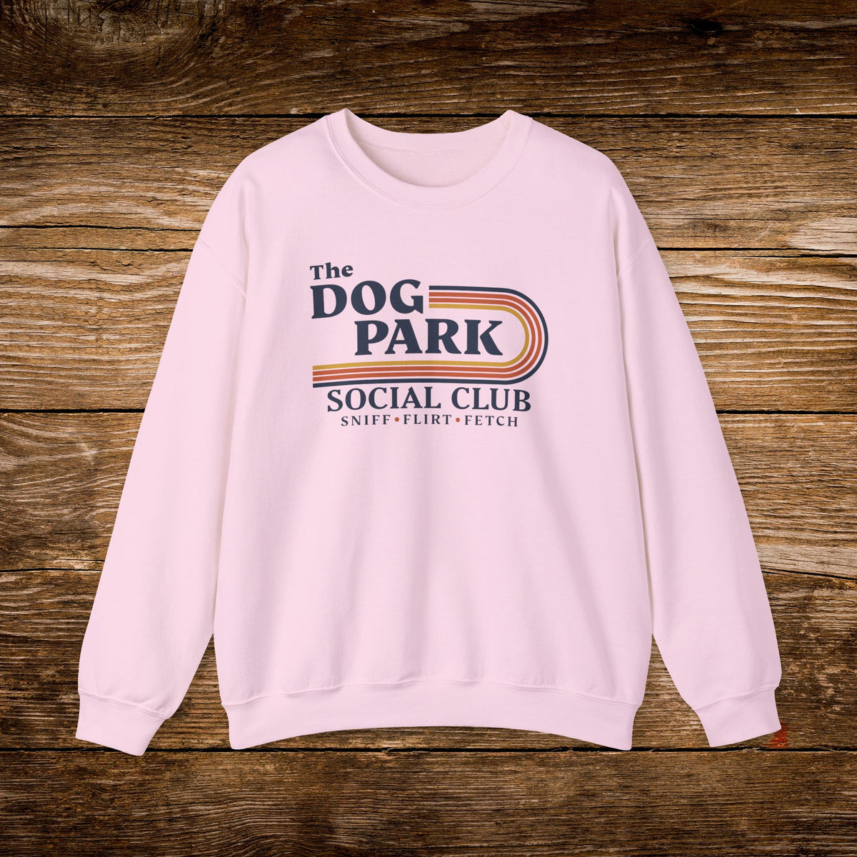 A light pink crewneck sweatshirt with a retro-style graphic reading 'The DOG PARK SOCIAL CLUB' and 'SNIFF - FLIRT - FETCH' in navy and red, displayed flat against a rustic brown wooden plank background.