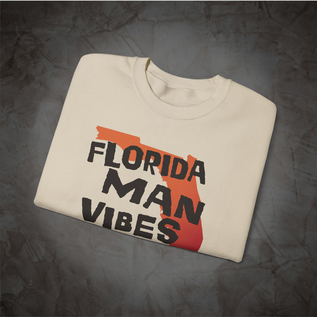A folded cream-colored t-shirt with a graphic print featuring the text 'FLORIDA MAN VIBES' in black over an orange and red map of Florida, displayed on a dark textured background.