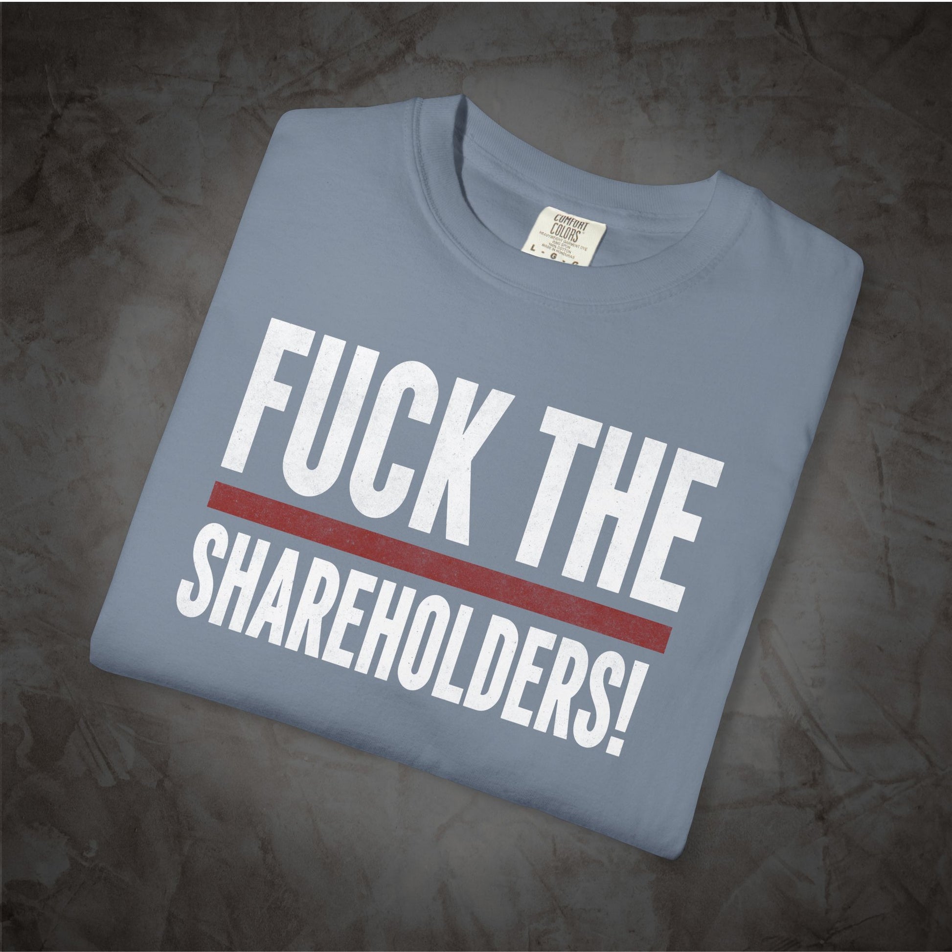 A folded light blue t-shirt with the text 'FUCK THE SHAREHOLDERS!' printed in bold white letters and a red horizontal stripe, displayed on a dark textured background.