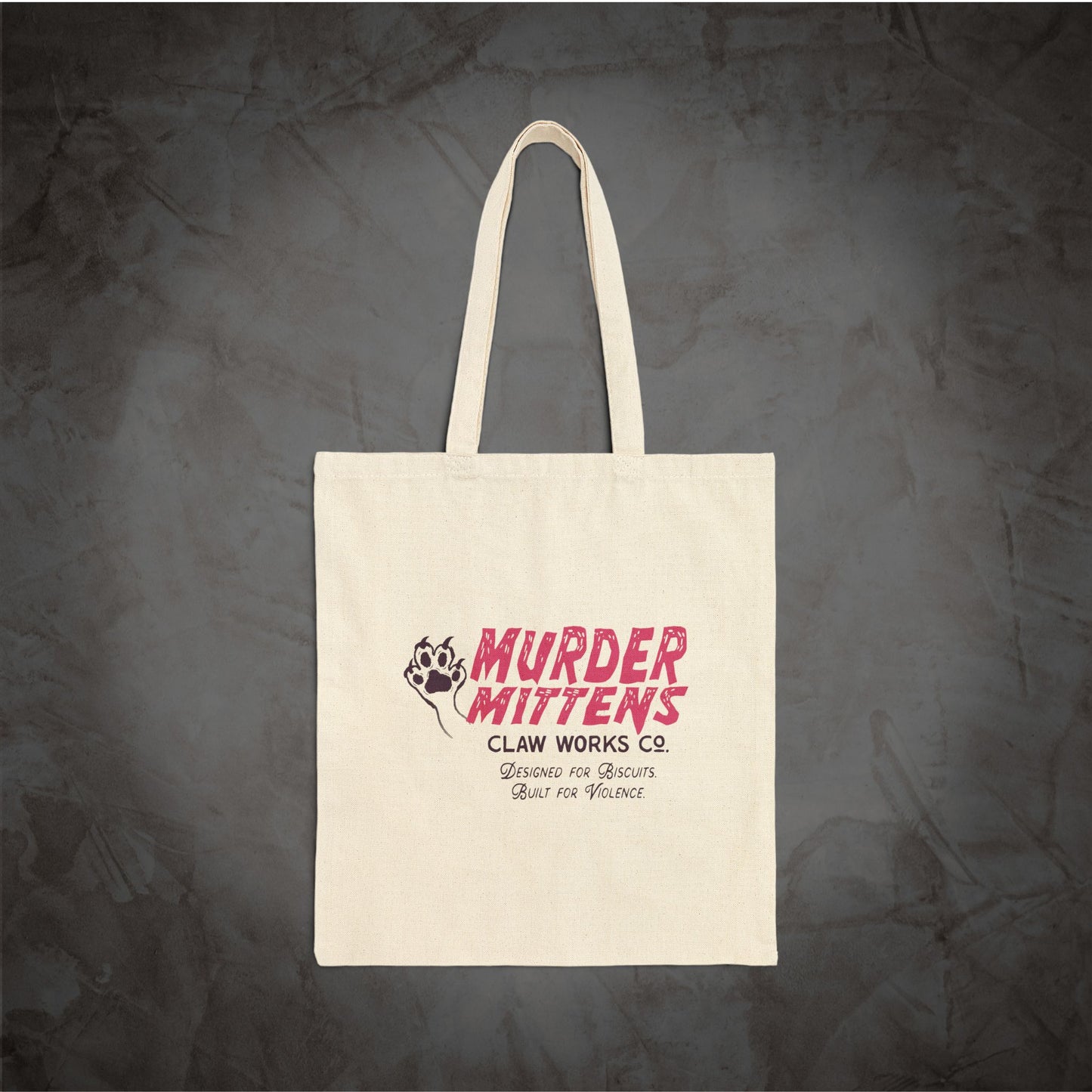 An off-white canvas tote bag with long handles, featuring a pink and black graphic print that reads 'MURDER MITTENS CLAW WORKS CO.' against a dark textured gray background.