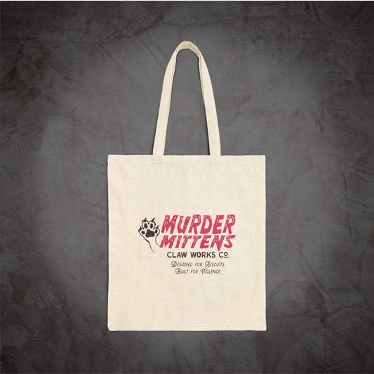 An off-white canvas tote bag with long handles, featuring a pink and black graphic print that reads 'MURDER MITTENS CLAW WORKS CO.' against a dark textured gray background.