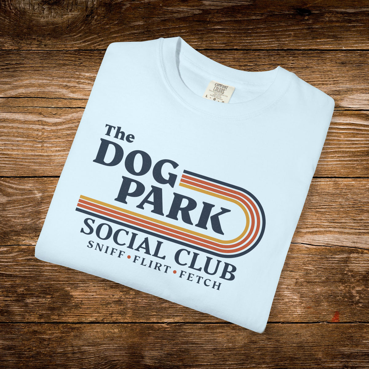 A folded light blue t-shirt with a retro-style graphic reading 'The DOG PARK SOCIAL CLUB' and 'SNIFF • FLIRT • FETCH' in dark blue and orange-yellow stripes, displayed on a rustic wooden background.