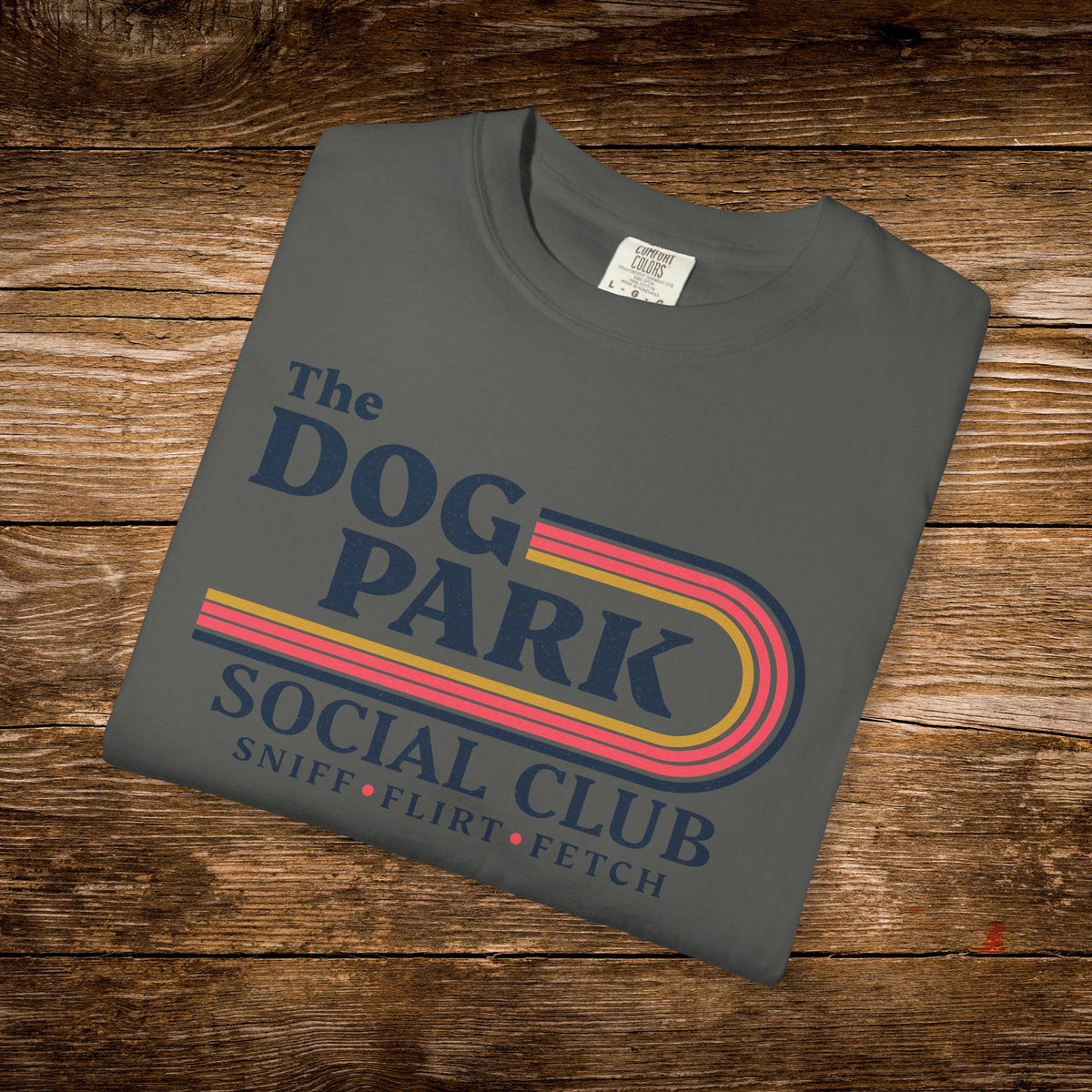 A dark gray folded t-shirt with a retro-style graphic reading 'The DOG PARK SOCIAL CLUB' and 'SNIFF • FLIRT • FETCH' in navy, pink, yellow, and orange, displayed on a rustic wooden background.