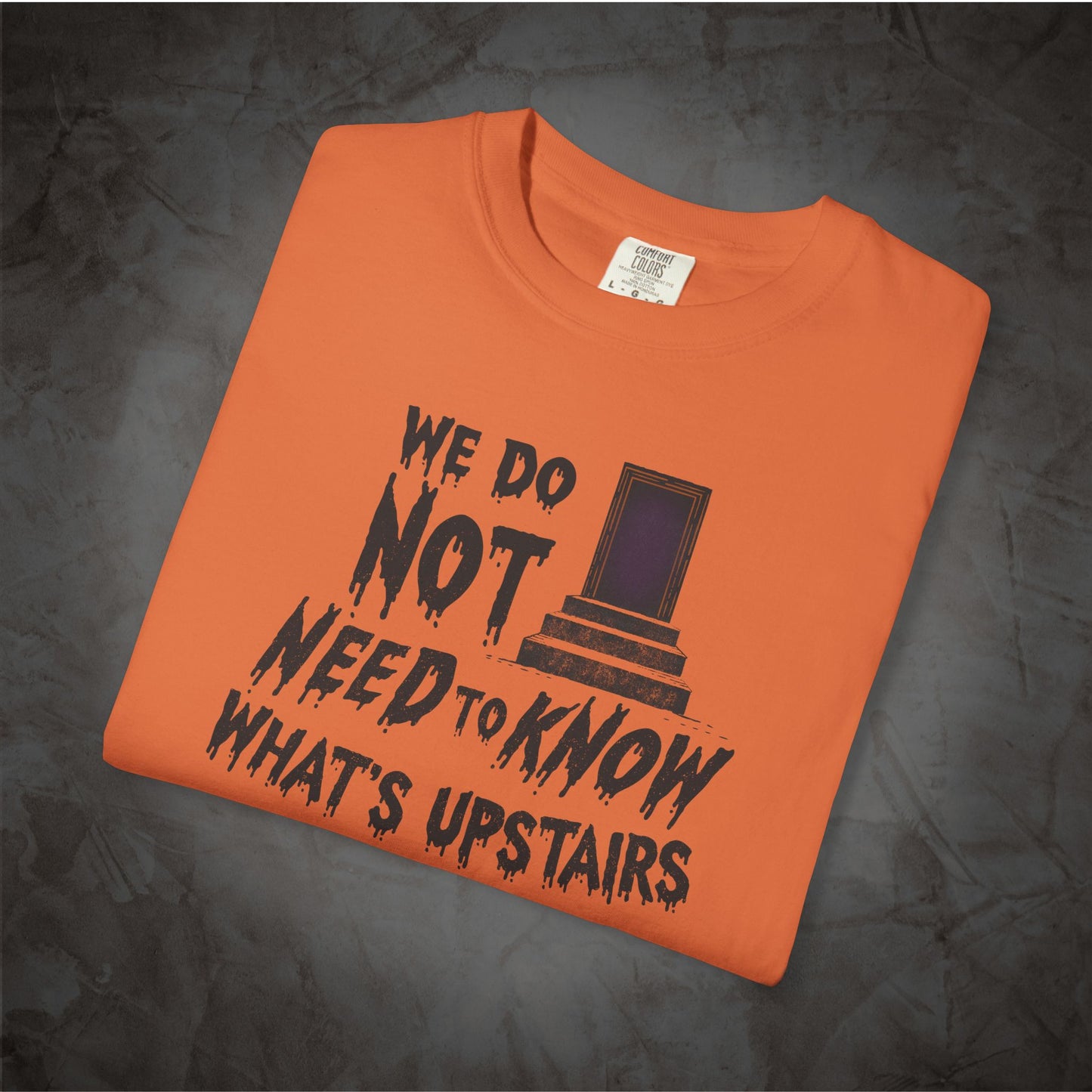 An orange folded t-shirt with a black graphic print that reads 'WE DO NOT NEED TO KNOW WHAT'S UPSTAIRS' alongside a depiction of the TARDIS from Doctor Who, displayed on a dark textured background.