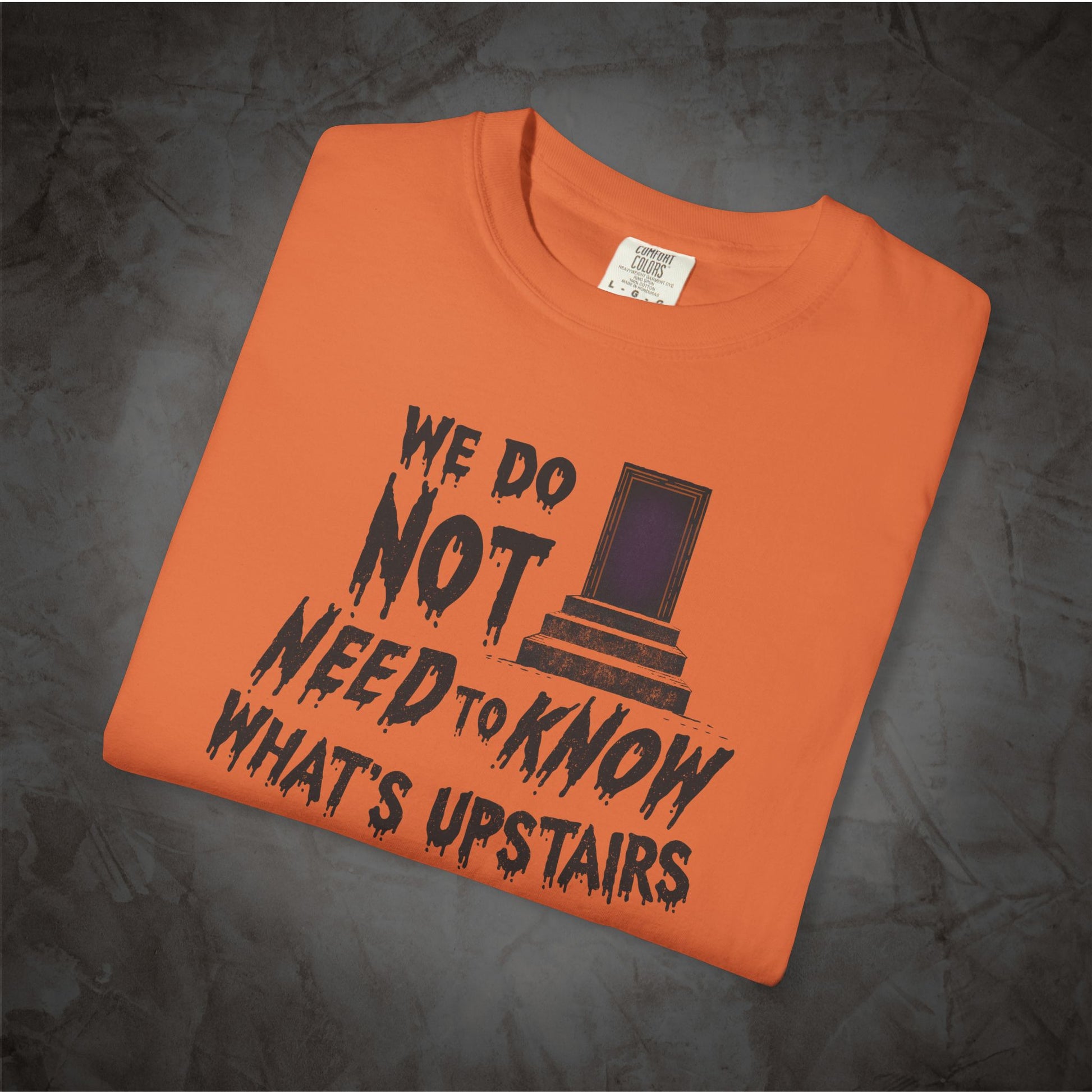 An orange folded t-shirt with a black graphic print that reads 'WE DO NOT NEED TO KNOW WHAT'S UPSTAIRS' alongside a depiction of the TARDIS from Doctor Who, displayed on a dark textured background.