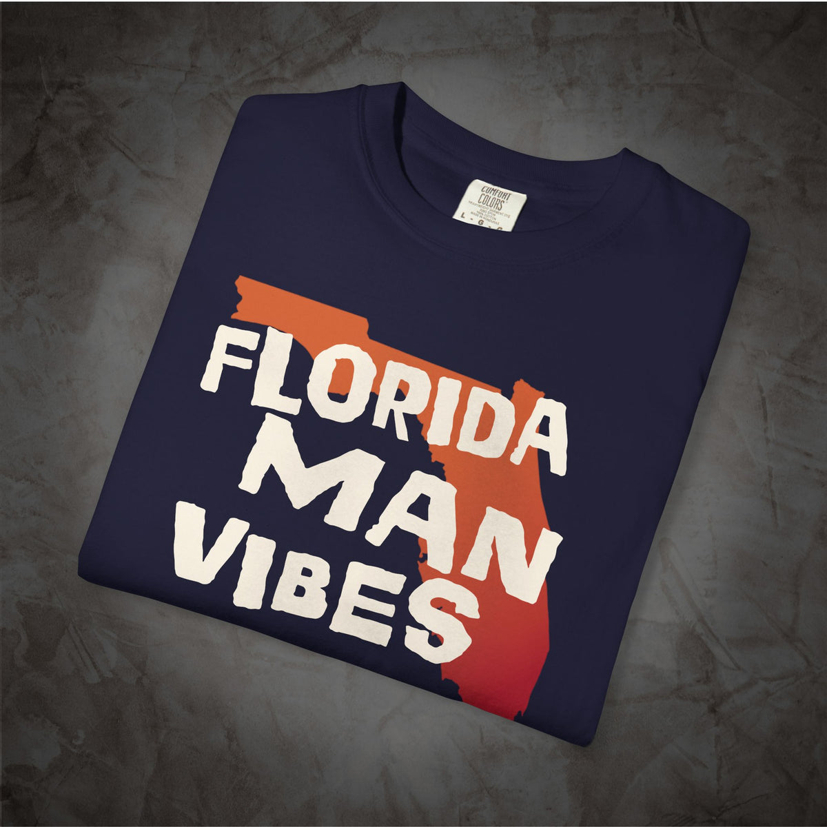 A navy blue folded t-shirt with a graphic print that reads 'FLORIDA MAN VIBES' in white text over an orange and red Florida state silhouette, displayed on a textured gray background.