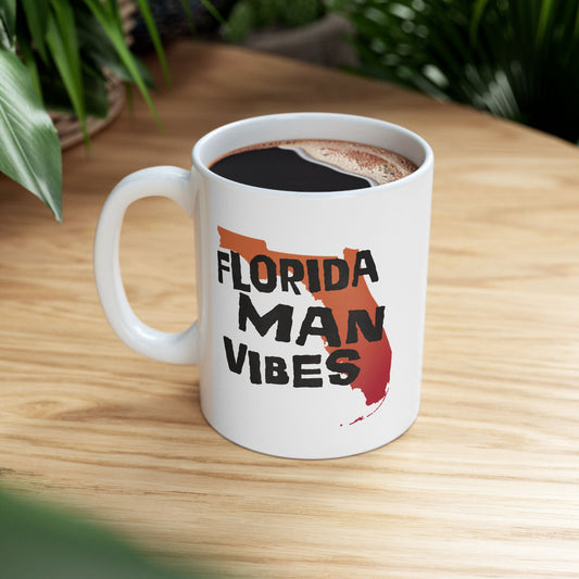 A white ceramic coffee mug with a black handle, filled with black coffee, sitting on a light wood table. The mug features a graphic design of the state of Florida filled with a red-orange gradient and the text 'FLORIDA MAN VIBES' in black.