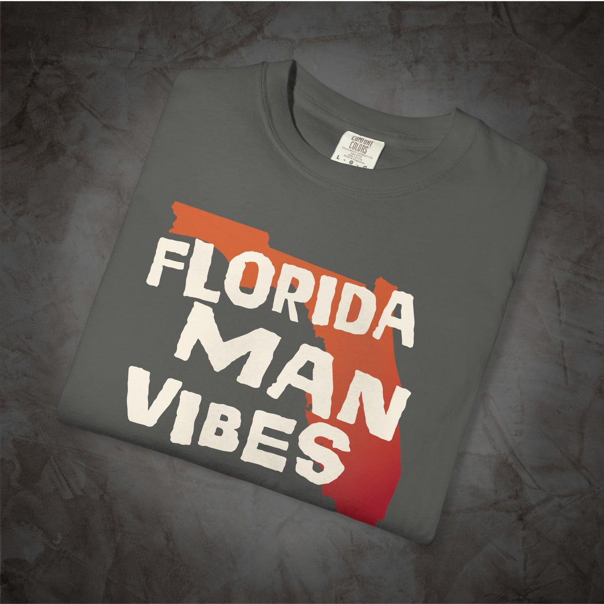 A folded dark gray t-shirt with a graphic print that reads 'FLORIDA MAN VIBES' in white text over a red and orange Florida state silhouette, displayed on a textured gray background.