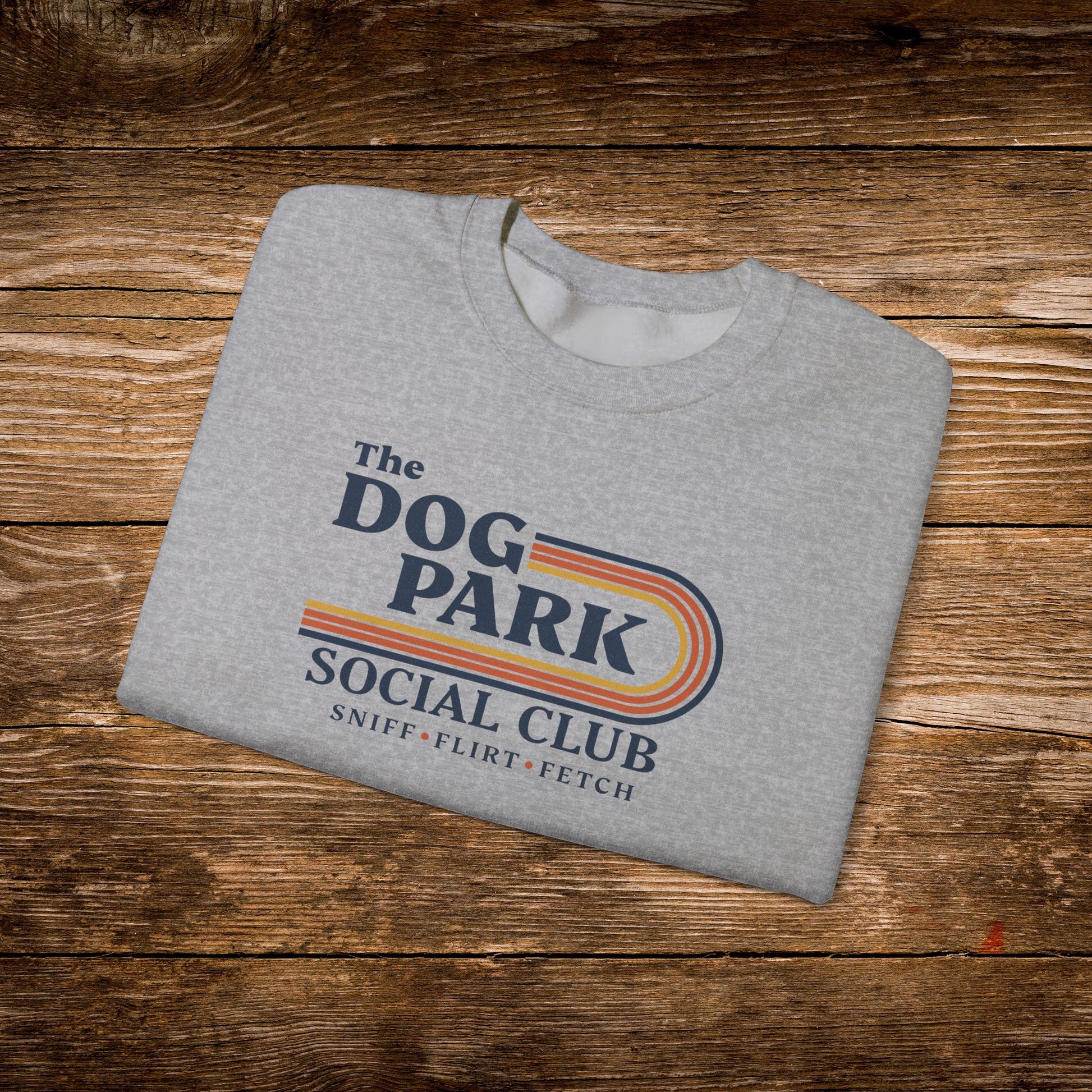 A folded heather gray t-shirt with a retro-style graphic reading 'The Dog Park Social Club' and 'Sniff, Flirt, Fetch' in navy and orange-yellow stripes, displayed on a rustic wooden plank background.