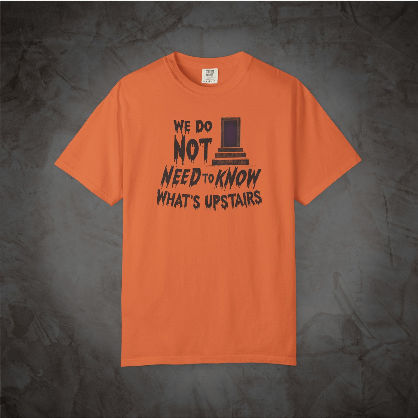 An orange short-sleeve t-shirt with a black graphic print on the front reading 'WE DO NOT NEED TO KNOW WHAT'S UPSTAIRS' alongside a top hat illustration, displayed against a dark textured background.