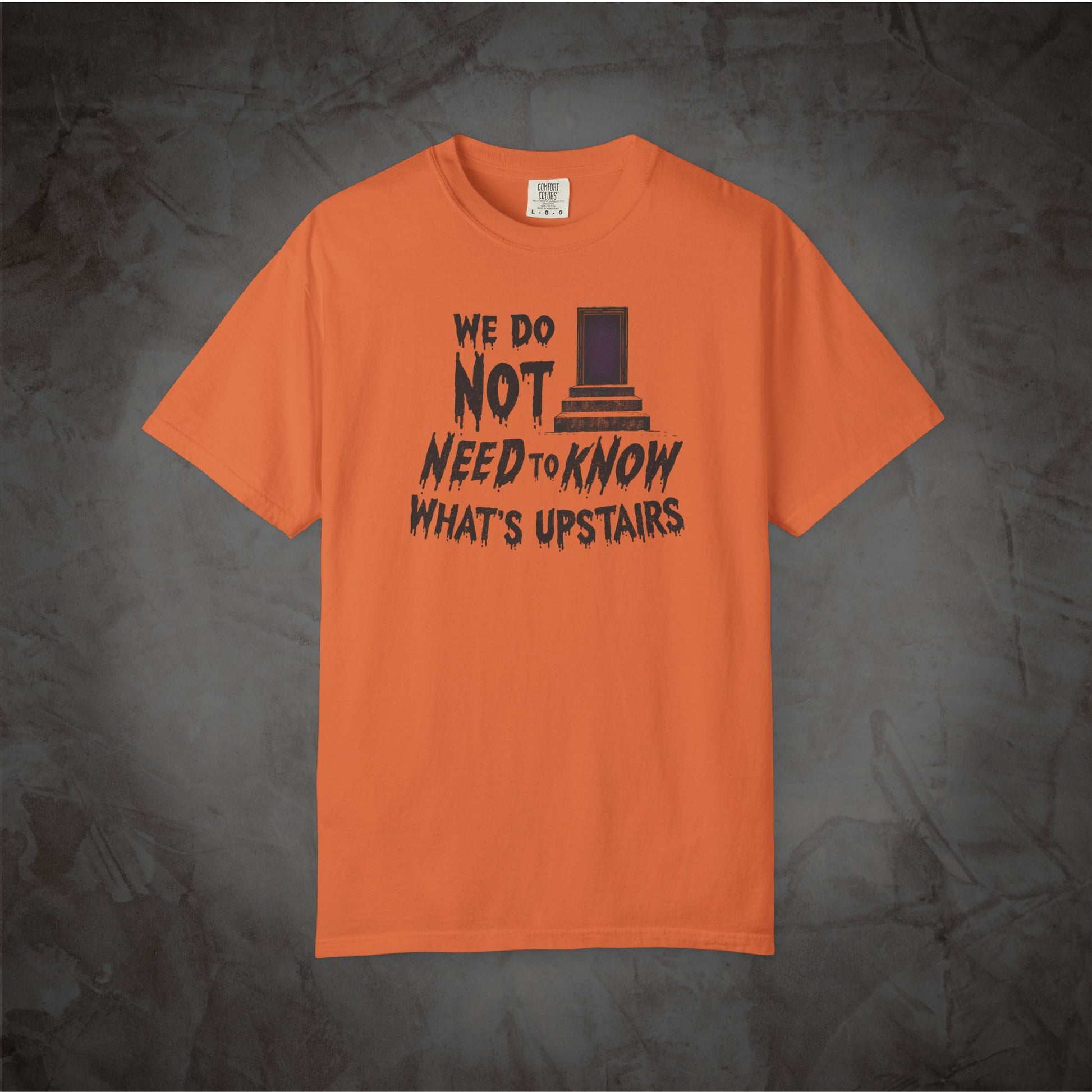 An orange short-sleeve t-shirt with a black graphic print on the front reading 'WE DO NOT NEED TO KNOW WHAT'S UPSTAIRS' alongside a top hat illustration, displayed against a dark textured background.