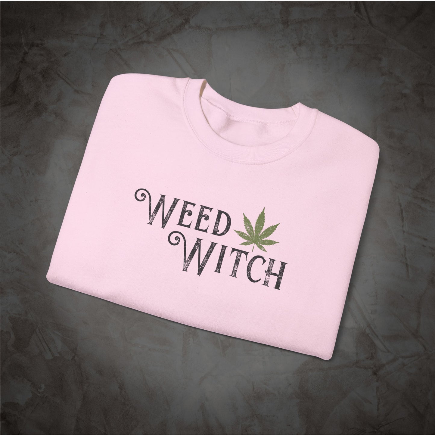 A light pink folded crewneck sweatshirt with 'WEED WITCH' printed in black gothic-style lettering and a green cannabis leaf graphic, displayed on a dark textured background.