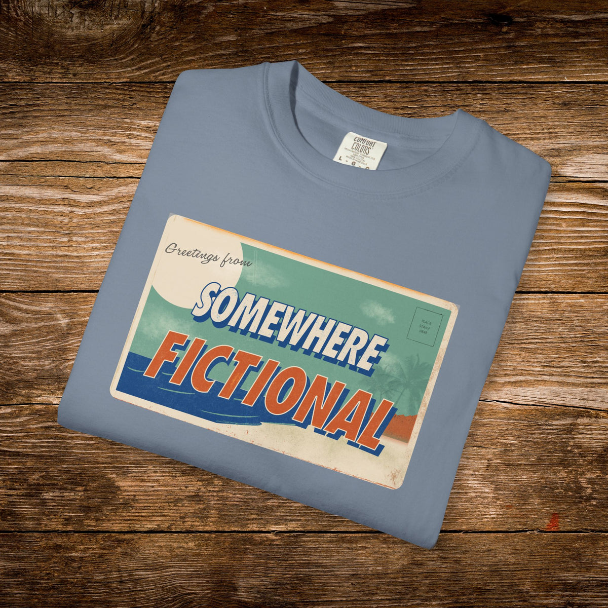 Folded Blue Jean Comfort Colors t-shirt featuring the “Greetings from Somewhere Fictional” postcard illustration.