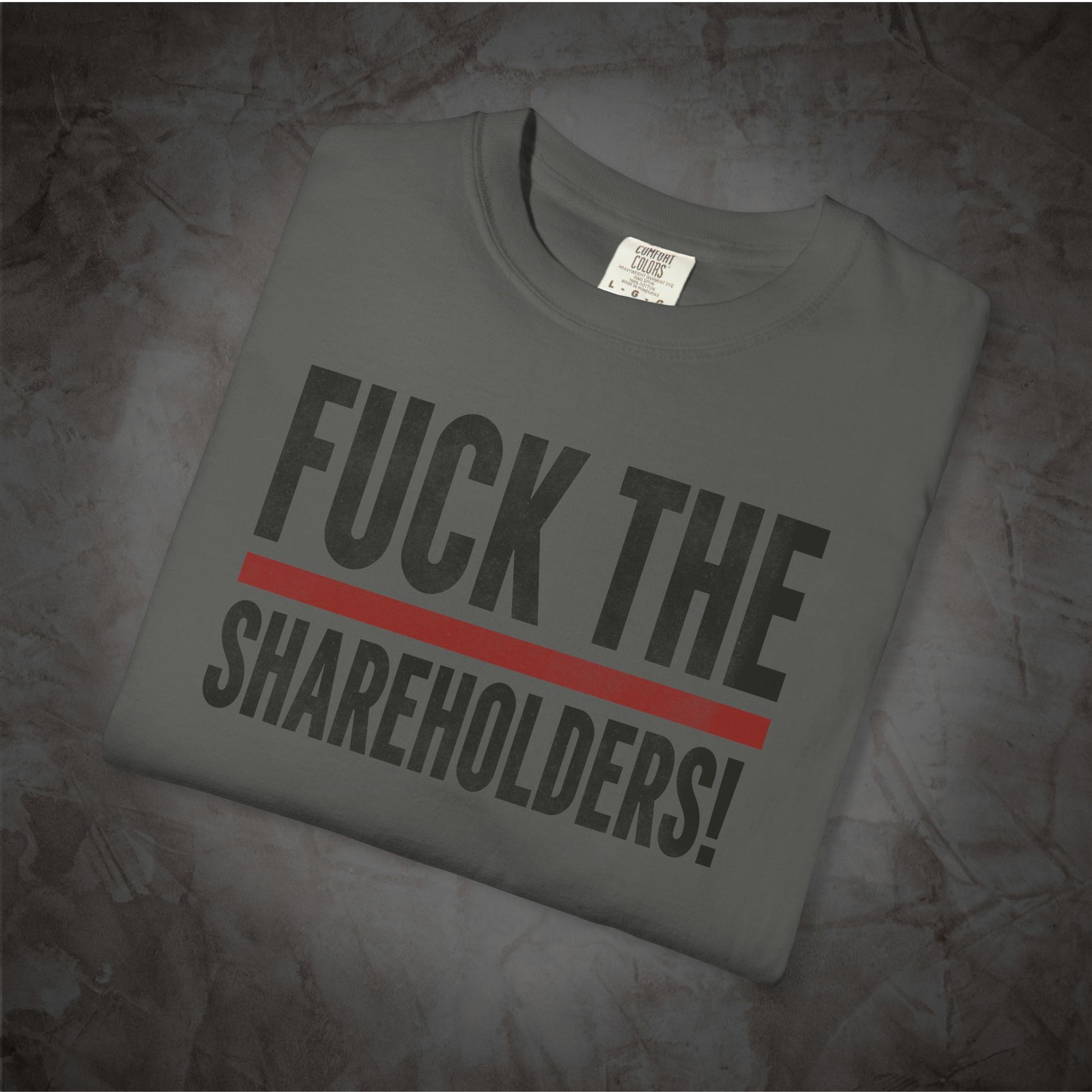 A folded dark gray t-shirt with the text 'FUCK THE SHAREHOLDERS!' printed in bold black letters and a red horizontal stripe, displayed on a textured gray background.