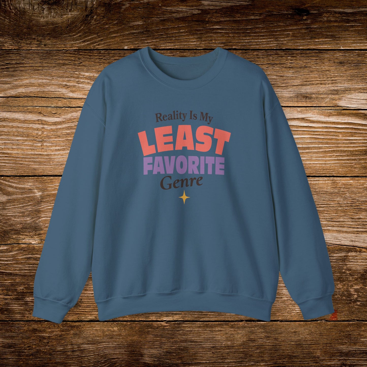 A slate blue crewneck sweatshirt with a colorful graphic text print reading 'Reality Is My LEAST FAVORITE Genre' on a rustic wooden background.