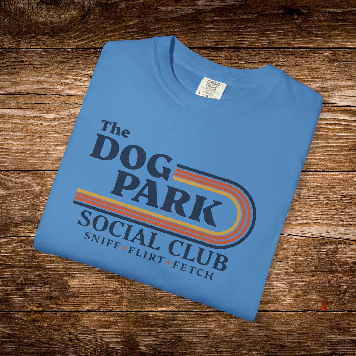 A folded light blue t-shirt with a retro-style graphic reading 'The DOG PARK SOCIAL CLUB' and 'SNIFF • FLIRT • FETCH' in dark blue text with orange, yellow, and red stripes, displayed on a rustic wooden background.