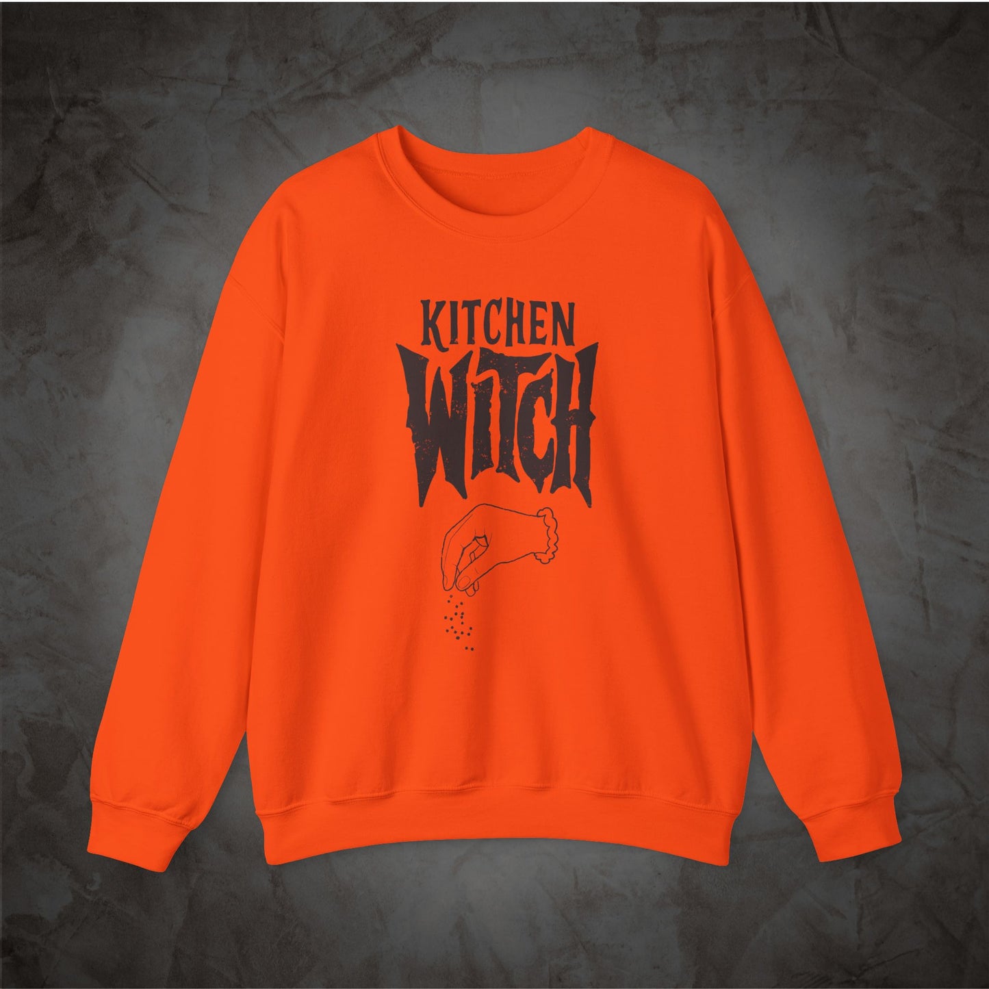 A bright orange crewneck sweatshirt with a black graphic print reading 'KITCHEN WITCH' and a hand sprinkling spices, displayed on a dark textured background.