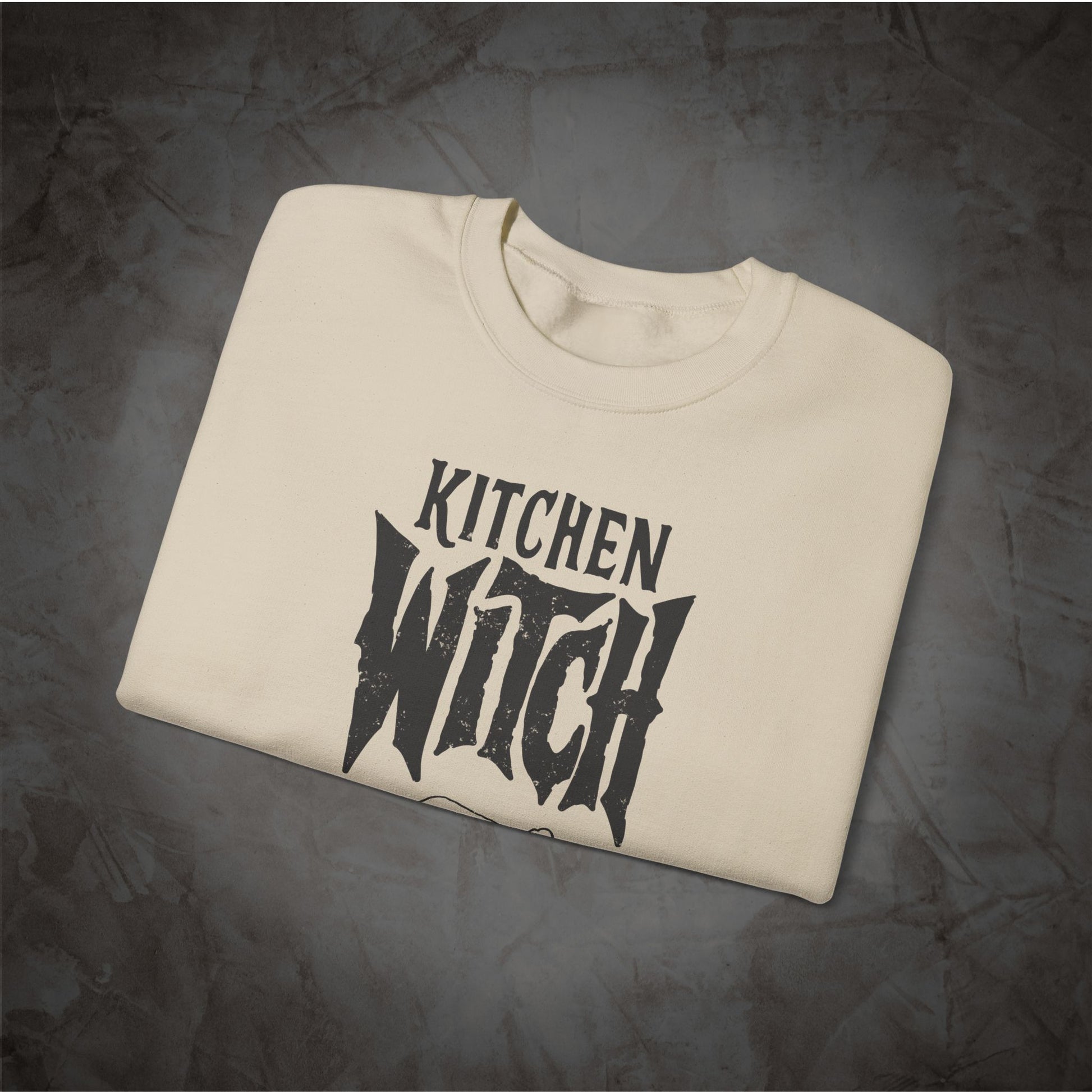 A cream-colored crewneck t-shirt with a black graphic reading 'KITCHEN WITCH' in a stylized font, folded neatly on a textured dark gray background.