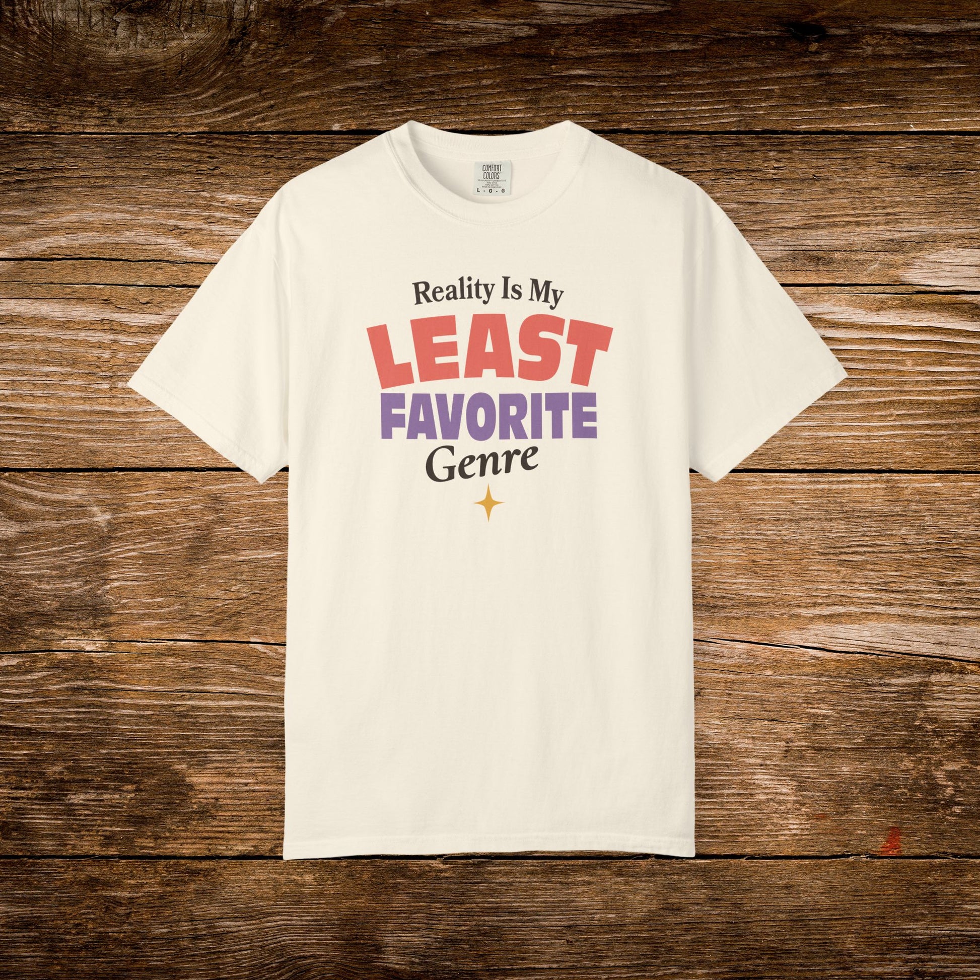An off-white short-sleeve t-shirt with a colorful graphic text print reading 'Reality Is My LEAST FAVORITE Genre' in red, purple, and black, displayed flat against a rustic brown wooden plank background.