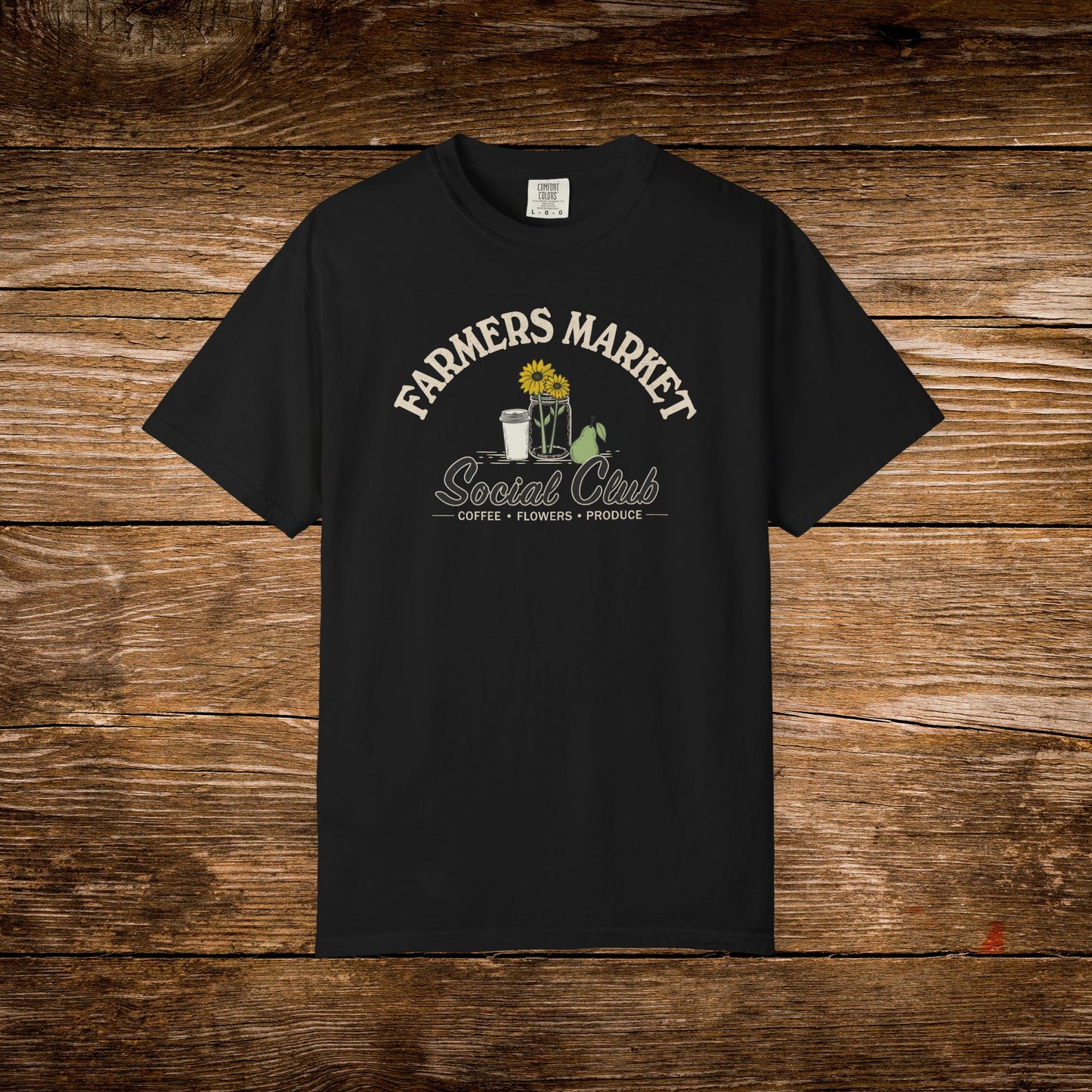 A black short-sleeve t-shirt with a vintage-style graphic on the chest reading 'FARMERS MARKET Social Club' in cream lettering, featuring a sunflower, coffee cup, flowers, and a watering can. The shirt is displayed flat against a rustic brown wooden plank background.