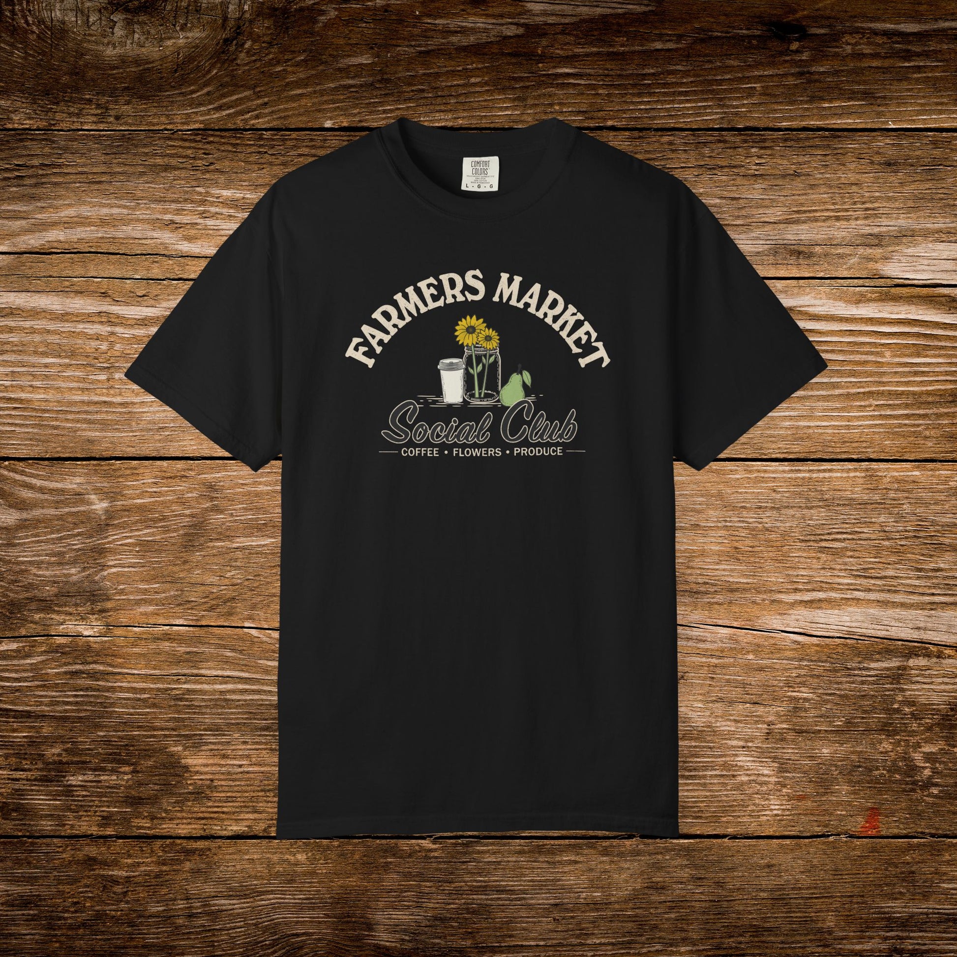 A black short-sleeve t-shirt with a vintage-style graphic on the chest reading 'FARMERS MARKET Social Club' in cream lettering, featuring a sunflower, coffee cup, flowers, and a watering can. The shirt is displayed flat against a rustic brown wooden plank background.