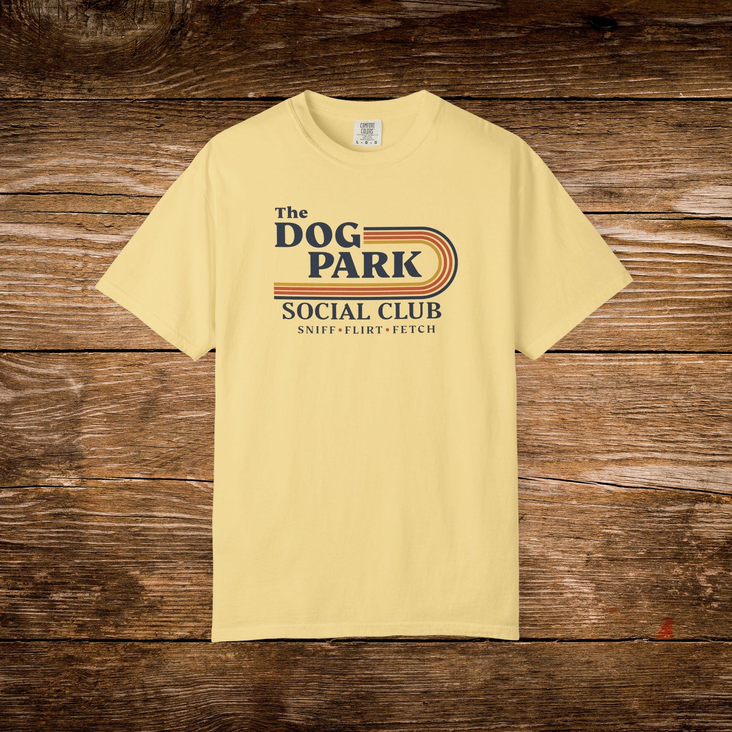 A pale yellow t-shirt with a retro-style graphic reading 'The DOG PARK SOCIAL CLUB' and 'SNIFF - FLEBT - FETCH' in navy and orange, displayed flat against a rustic dark wood background. The design text reads "Sniff, Flirt, Fetch."