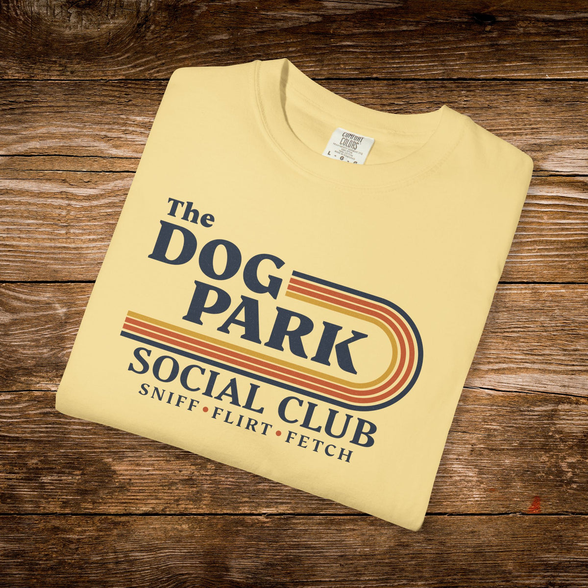 A folded cream-colored t-shirt with a retro-style graphic reading 'The DOG PARK SOCIAL CLUB' and 'SNIFF • FLIRT • FETCH' in navy and orange, displayed on a rustic wooden background.