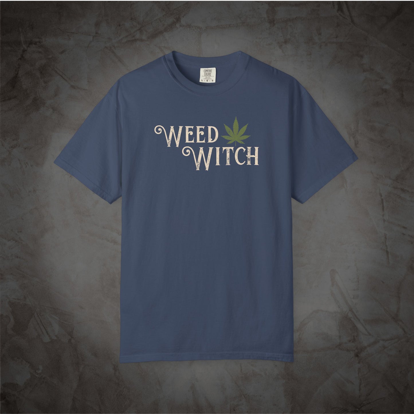 A slate blue short-sleeve t-shirt with a graphic on the chest reading 'WEED WITCH' in a cream-colored decorative font, featuring a green cannabis leaf as the letter 'O'. The shirt is displayed flat against a textured, mottled gray background.