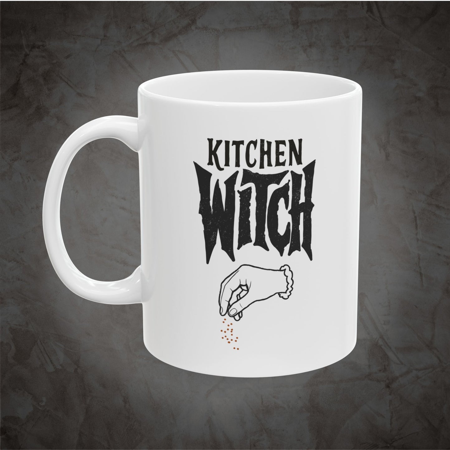 A white ceramic mug with a black graphic that reads 'KITCHEN WITCH' above a hand sprinkling small red dots, shown on a dark textured background.