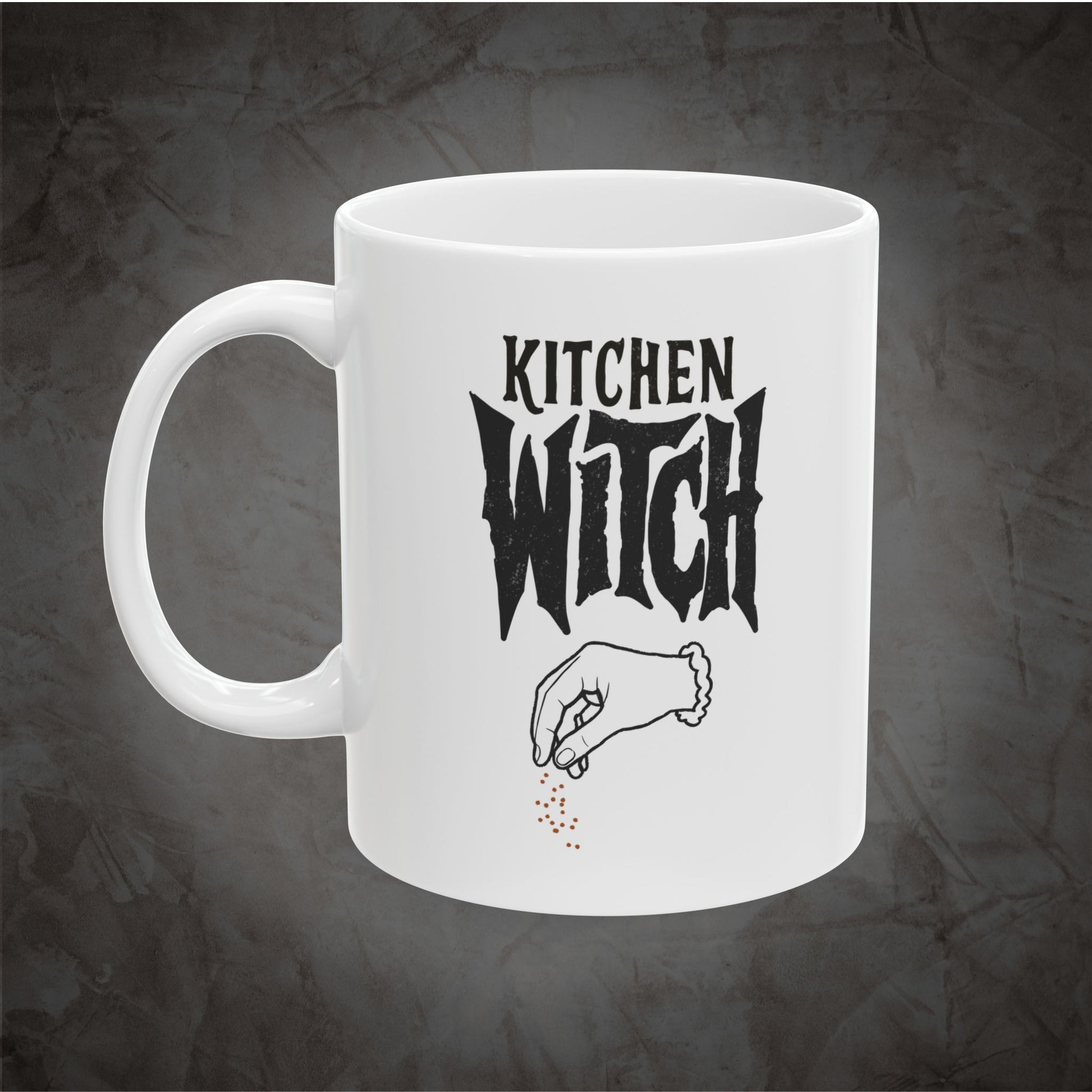 A white ceramic mug with a black graphic that reads 'KITCHEN WITCH' above a hand sprinkling small red dots, shown on a dark textured background.