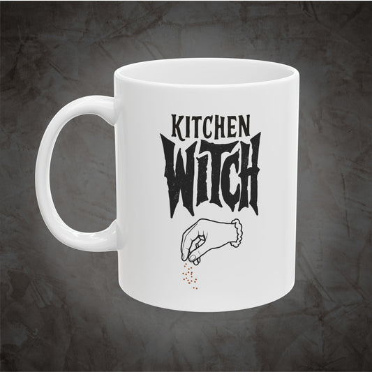 A white ceramic mug with a black graphic that reads 'KITCHEN WITCH' above a hand sprinkling small red dots, shown on a dark textured background.