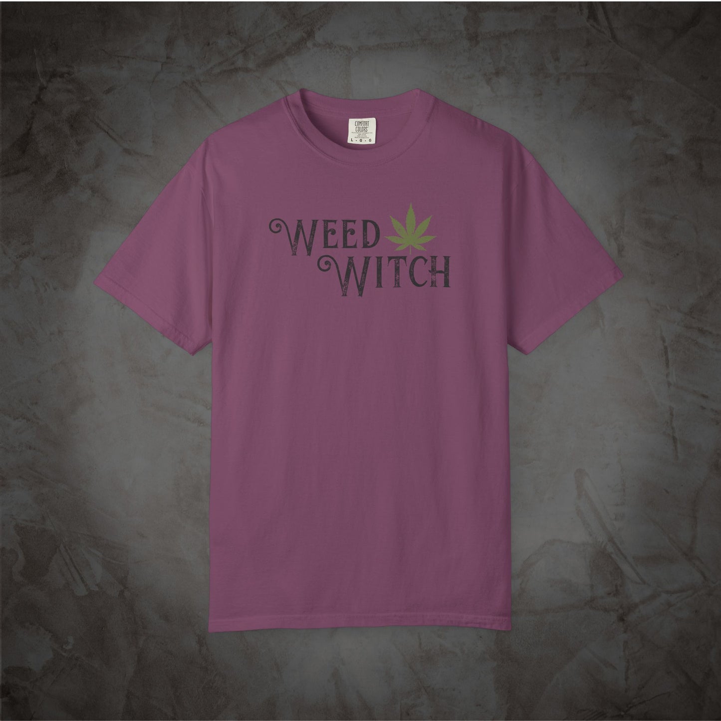 A mauve purple short-sleeve t-shirt with a 'WEED WITCH' graphic on the chest, featuring a green cannabis leaf, laid flat against a textured gray background.