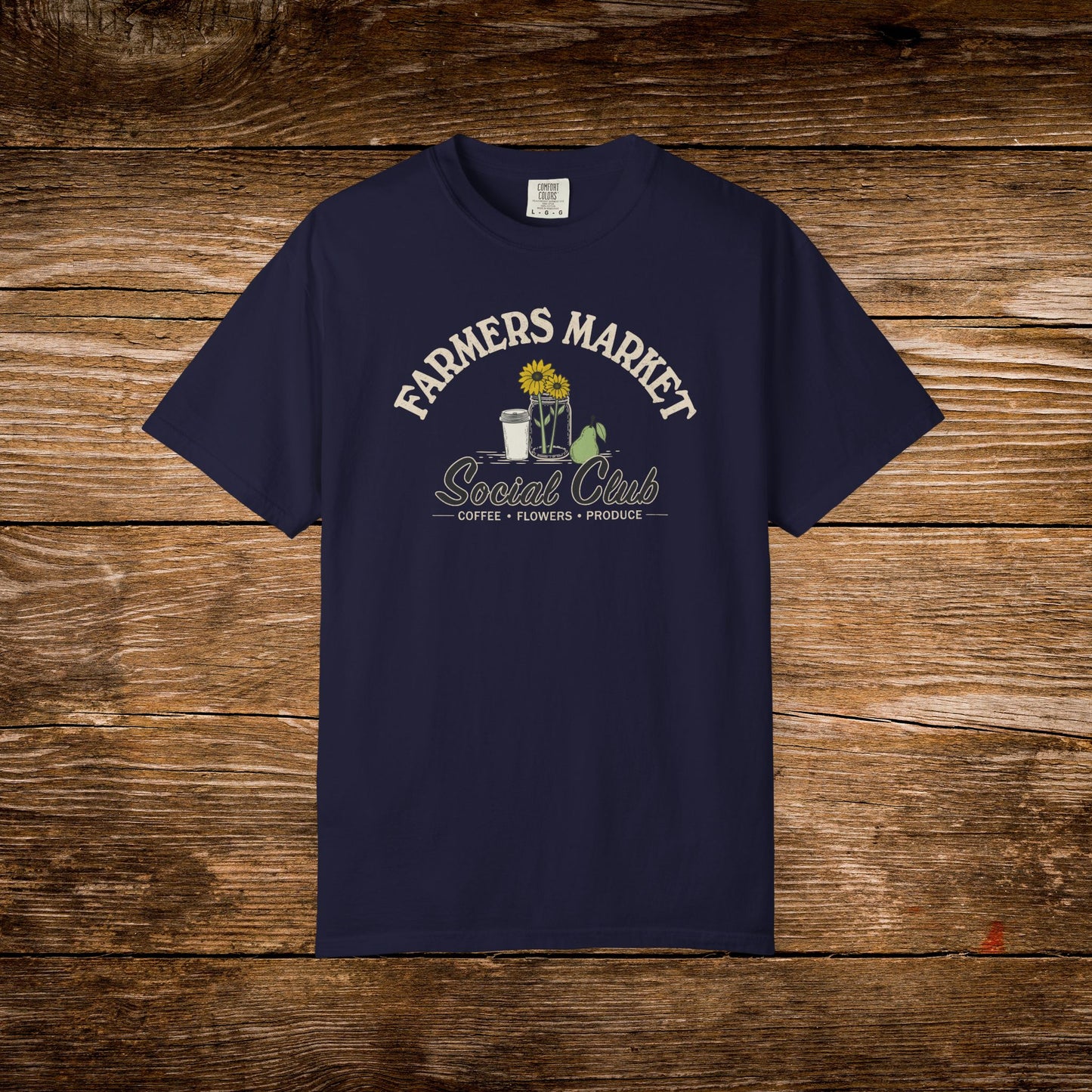 A navy blue short-sleeve t-shirt with a vintage-style graphic on the chest reading 'FARMERS MARKET Social Club' with a sunflower, coffee cup, flowers, and a watering can, displayed flat against a rustic brown wooden plank background.