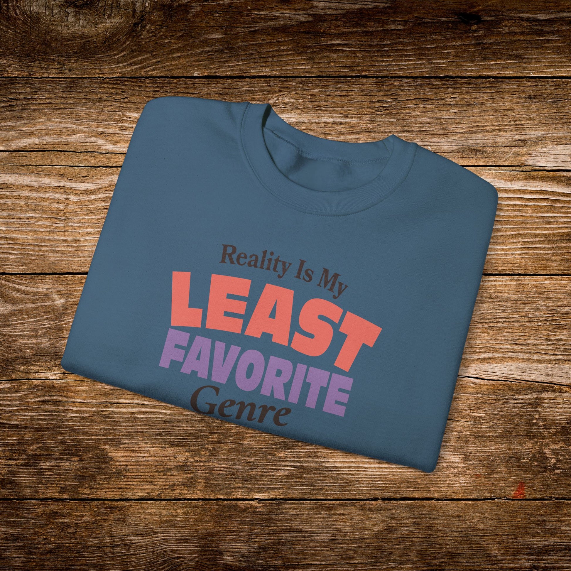 A folded slate blue t-shirt with the text 'Reality Is My LEAST FAVORITE Genre' printed on the front, displayed on a rustic wooden background.