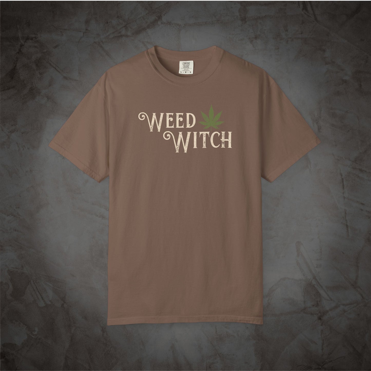 A brown short-sleeve t-shirt with 'WEED WITCH' printed in a light beige font on the chest, accompanied by a green cannabis leaf graphic, displayed against a dark textured gray background.