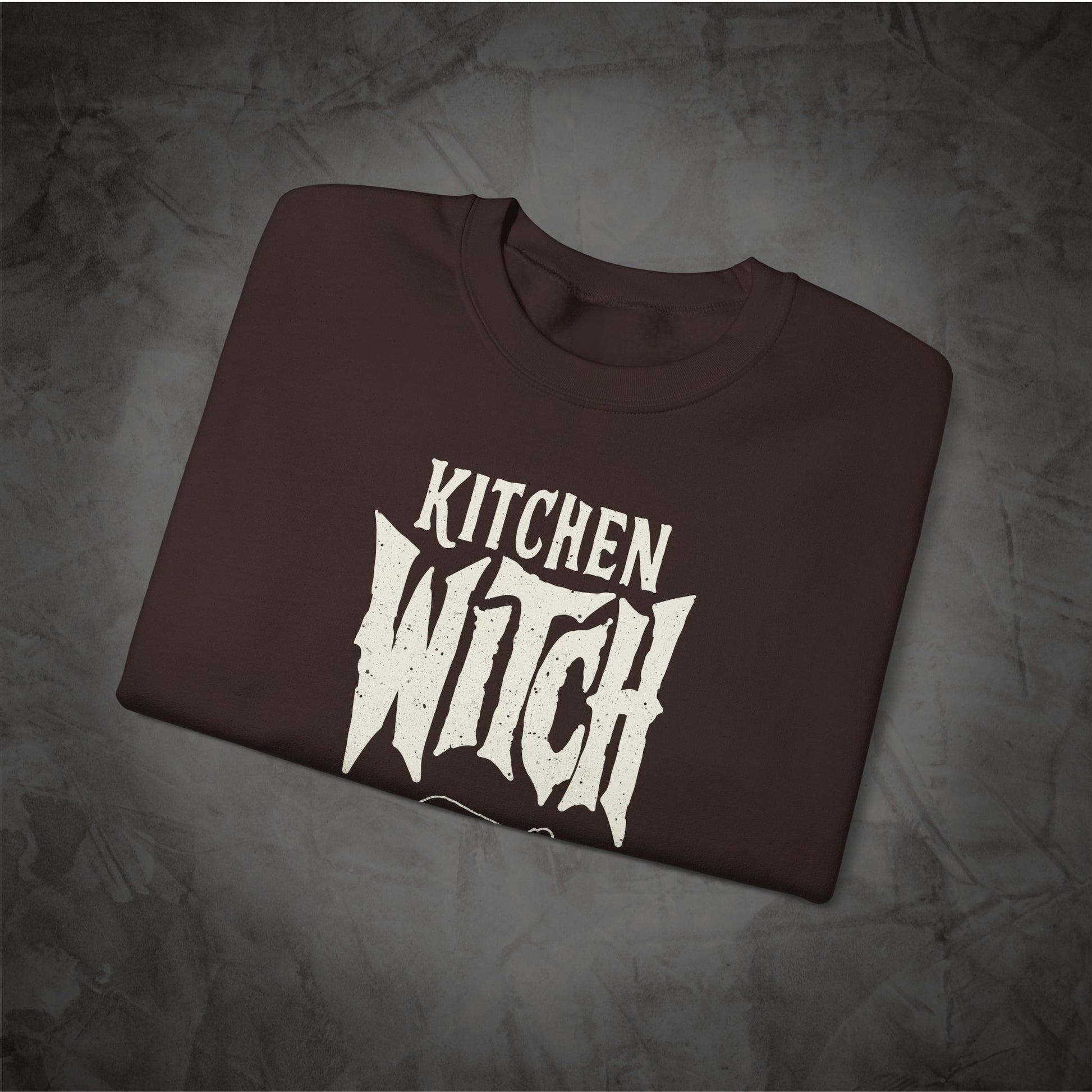 A dark brown folded t-shirt with a distressed white graphic reading 'KITCHEN WITCH' on a textured gray background.