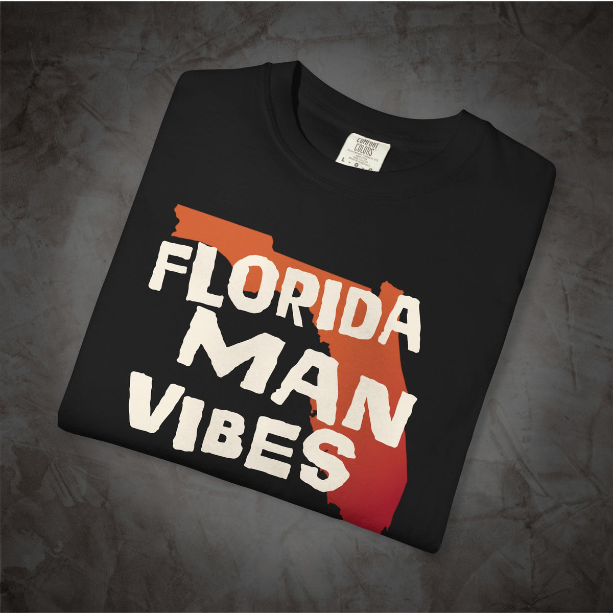 A folded black t-shirt with a graphic print that reads 'FLORIDA MAN VIBES' in white text over a red and orange Florida state silhouette, displayed on a textured gray background.