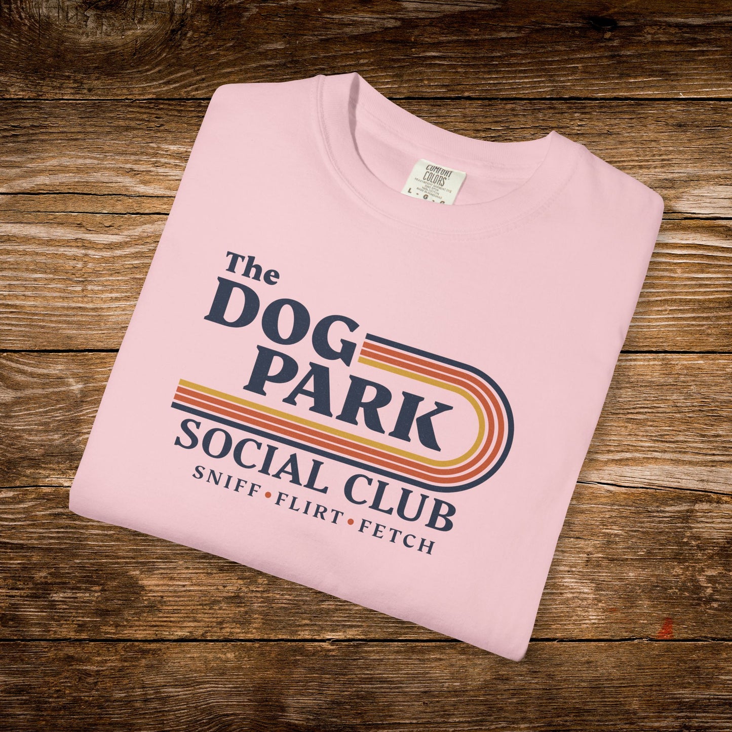 A folded light pink crewneck t-shirt with a retro-style graphic reading 'The DOG PARK SOCIAL CLUB' and 'SNIFF • FLIRT • FETCH' in navy, orange, and yellow, displayed on a rustic wooden background. The design text reads "Sniff, Flirt, Fetch."