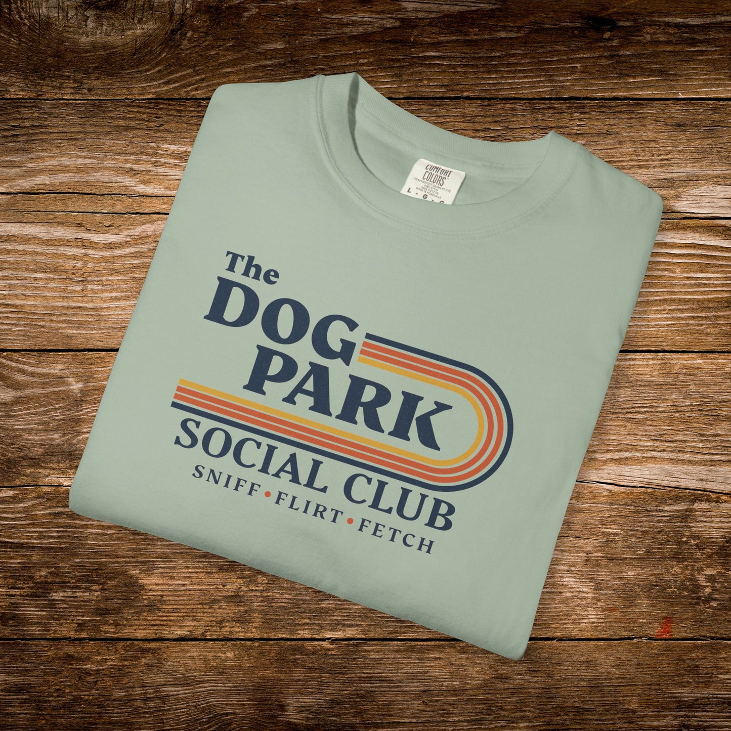 A folded sage green t-shirt with a retro-style graphic reading 'The DOG PARK SOCIAL CLUB' and 'SNIFF * FLIRT * FETCH' in navy, orange, and yellow, displayed on a rustic wooden background. The design text reads "Sniff, Flirt, Fetch."