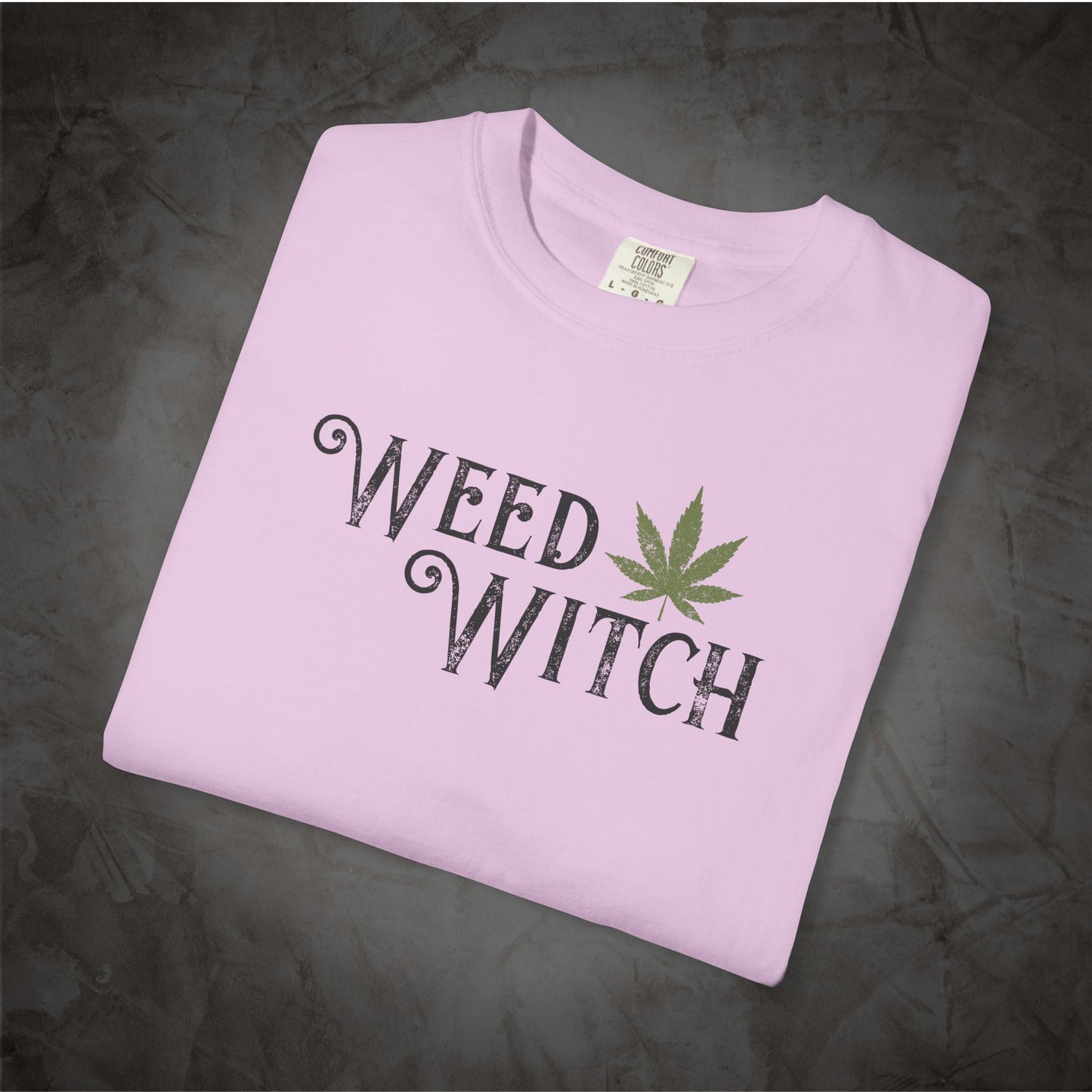 A light pink folded t-shirt with a 'WEED WITCH' graphic printed in black with a green cannabis leaf, displayed on a dark textured background.